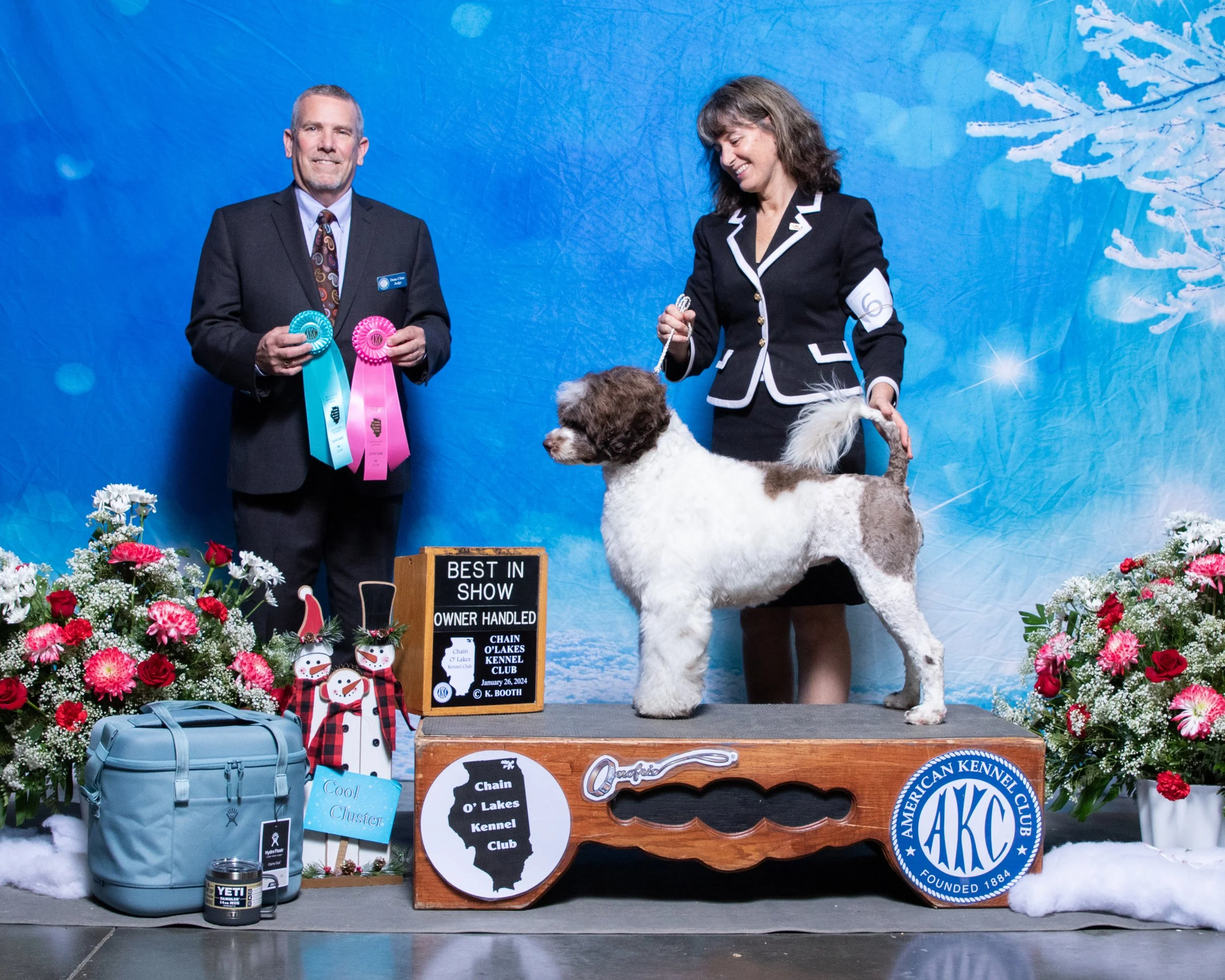 A dog show award ceremony with a brown and white dog on a platform, two people standing beside it holding ribbons, and decorated with winter-themed flowers and snowmen.