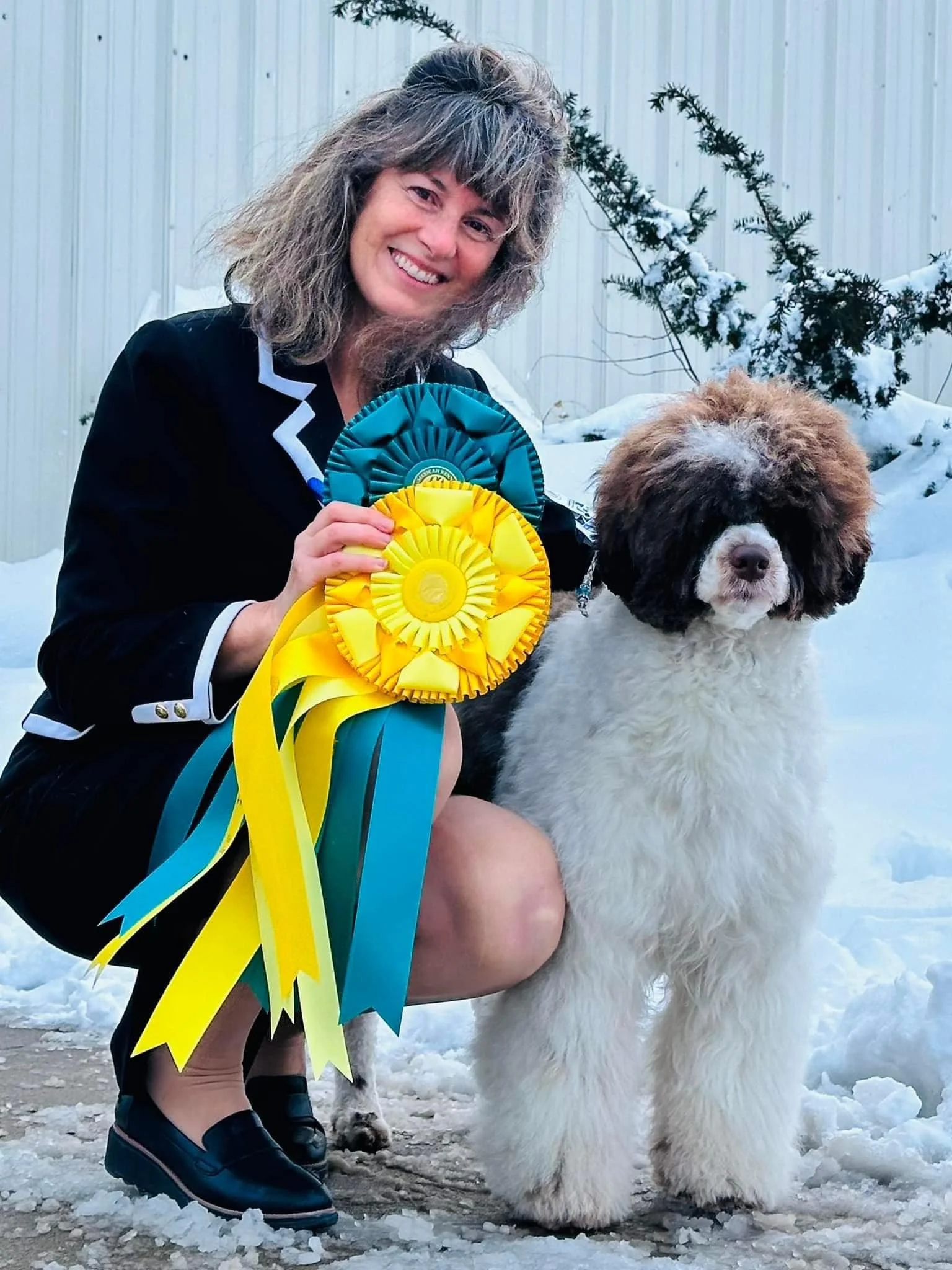 A woman with gray, curly hair kneeling on snow next to a large, fluffy dog with brown and white fur, holding two colorful ribbons, one blue and one yellow, in an outdoor winter setting.