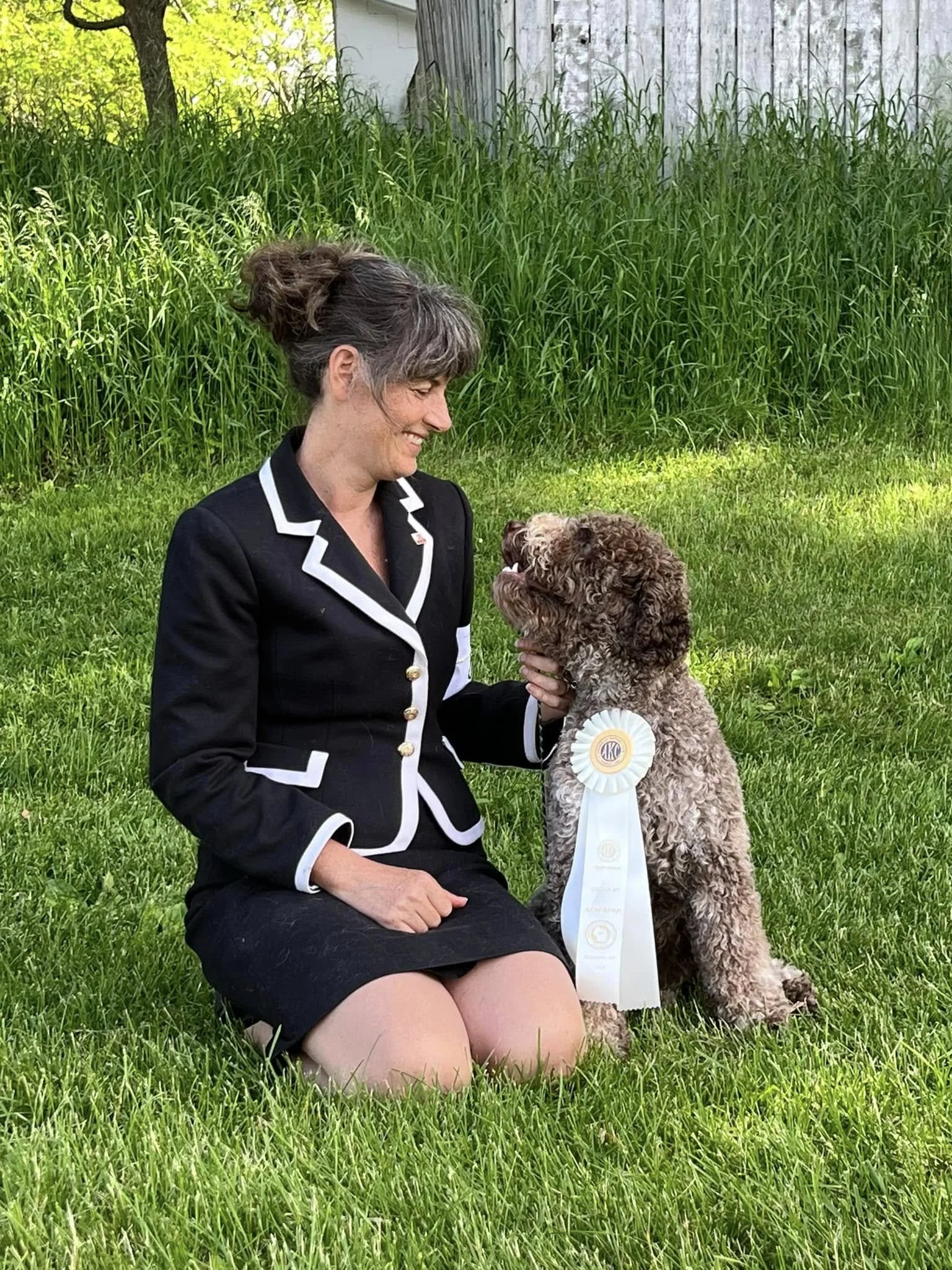 A woman kneeling on the grass with a curly-haired dog that has a large blue and white ribbon hanging from its neck, outdoors with tall grass and a tree in the background.