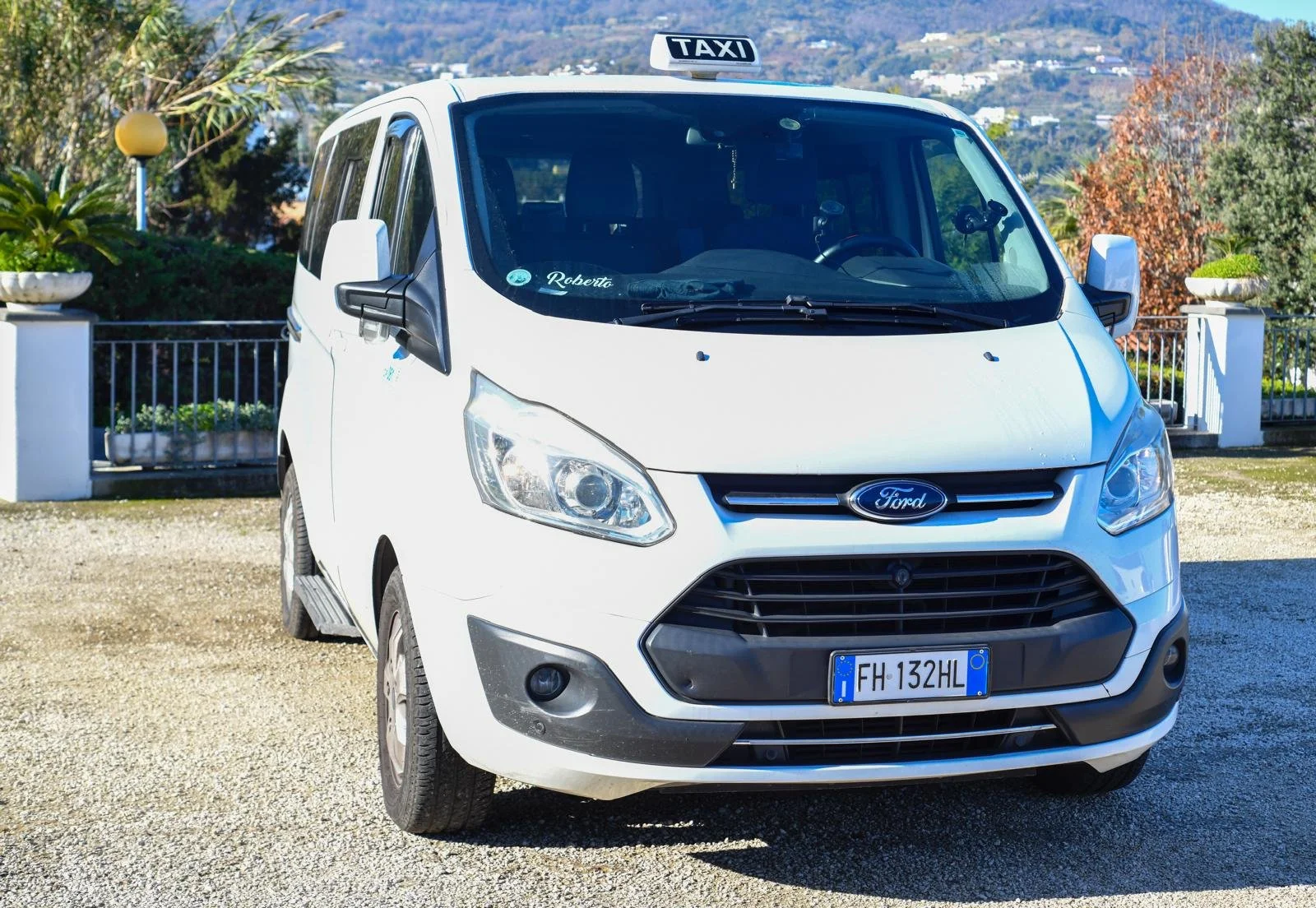Auto Ford bianca con targa FH 132HL e segnale taxi sul tetto, parcheggiata su una superficie di ghiaia davanti a un'area verde con piante e un cortile di pietra, sullo sfondo colline e case.