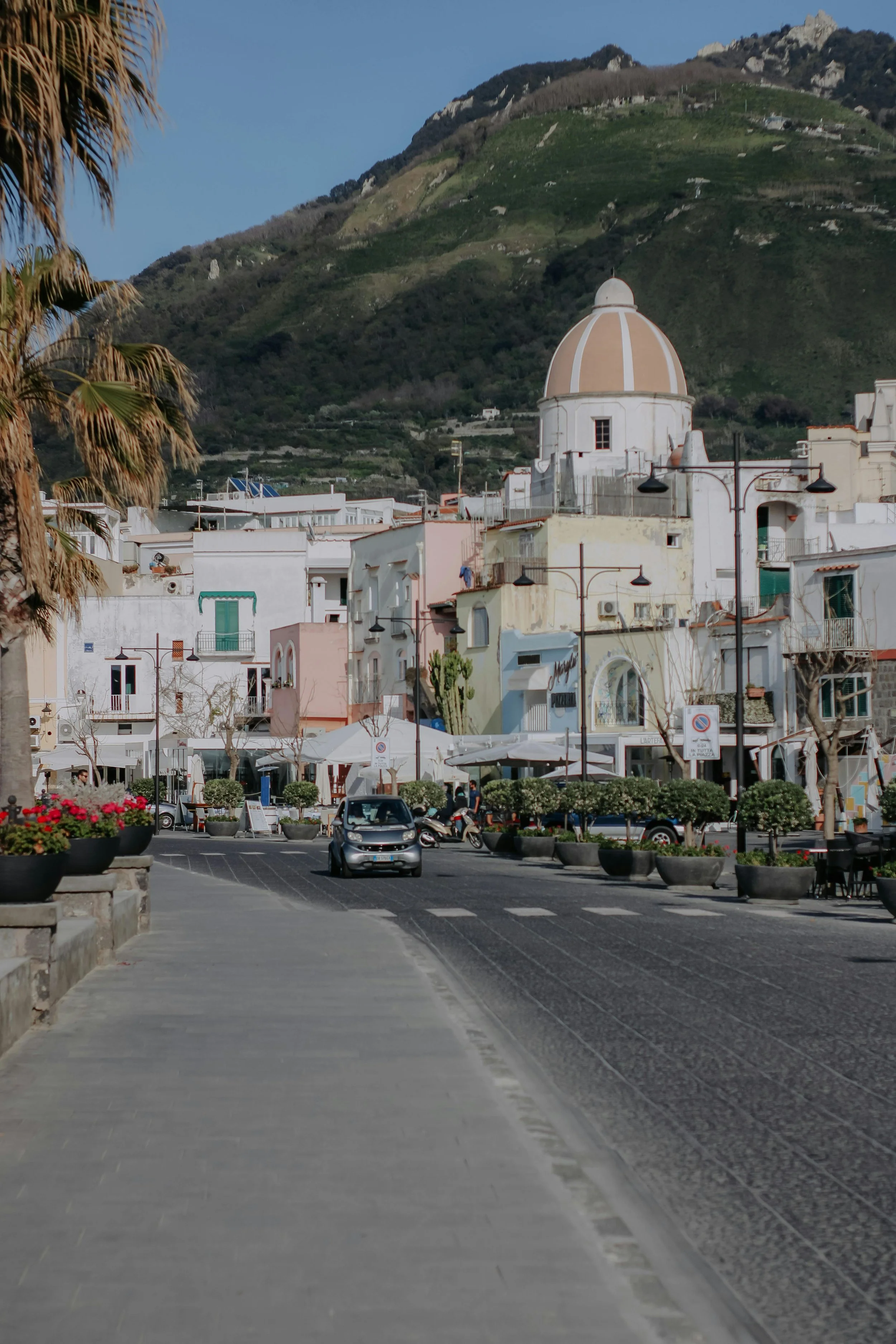 Veduta di una piazza di un paese mediterraneo con edifici bianchi, un'edicola con cupola, palme, piante ornamentali e montagne sullo sfondo.