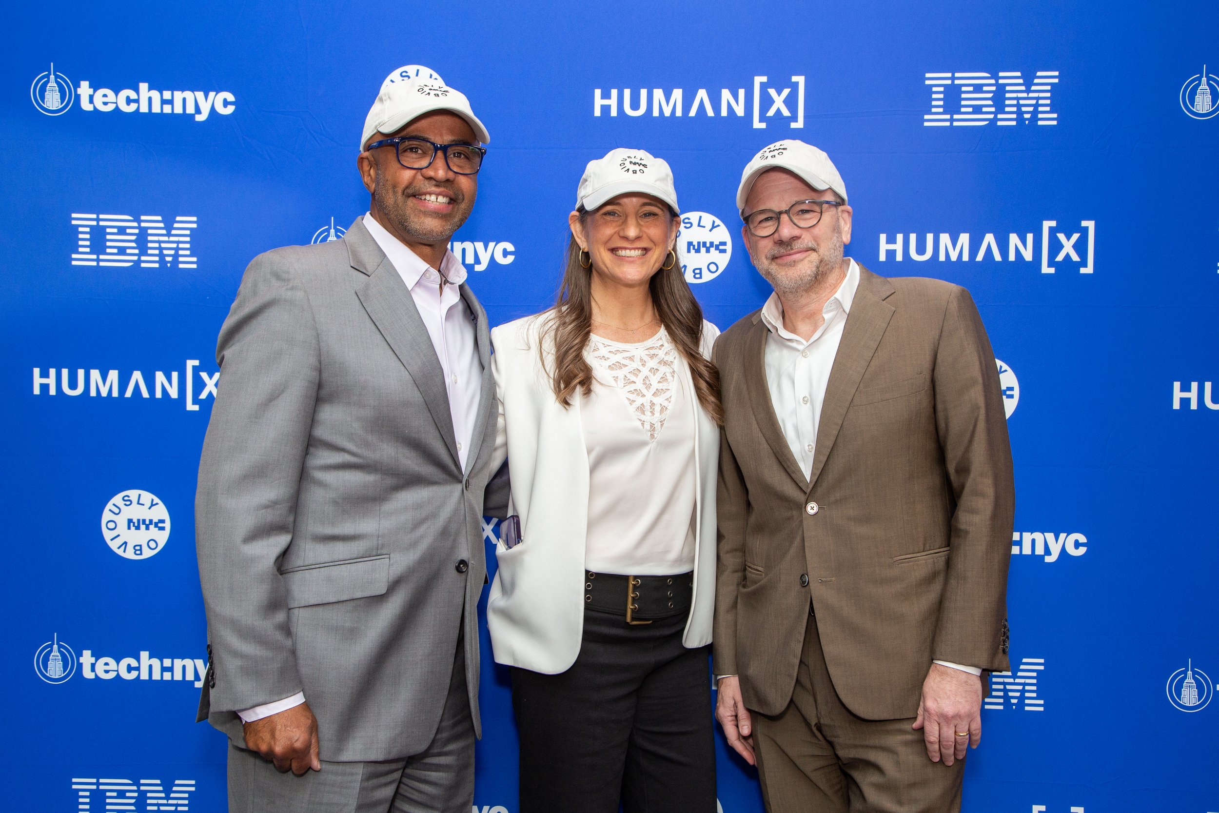 Three people standing in front of a blue backdrop with logos, wearing white caps. A man in a gray suit on the left, a woman in the middle with a white jacket and black pants, and a man in a brown suit on the right.