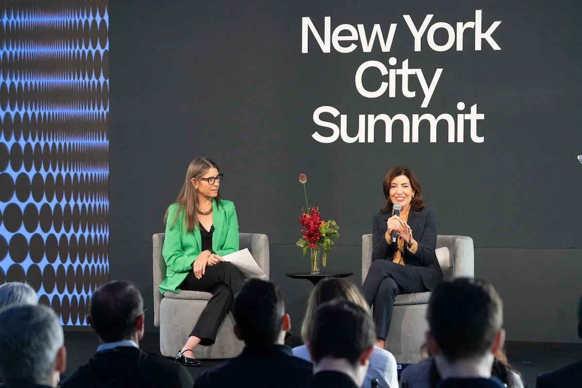 Two women speaking at the New York City Summit, seated on stage with a dark background and the event's name displayed. One woman is dressed in a black suit and holds a microphone; the other woman is dressed in a green blazer and is holding papers. Th