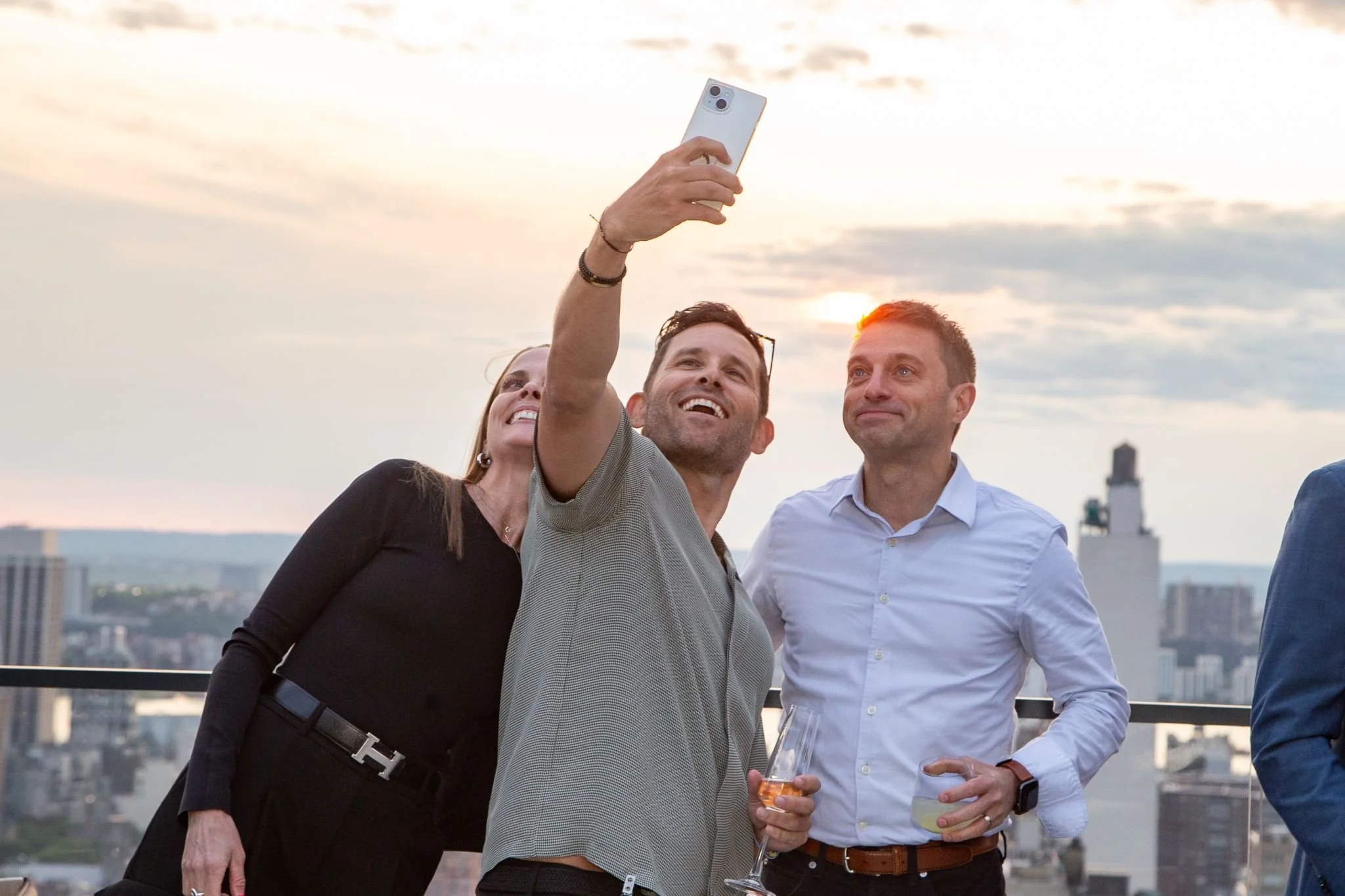 People taking a selfie on a rooftop during sunset with a city skyline in the background.