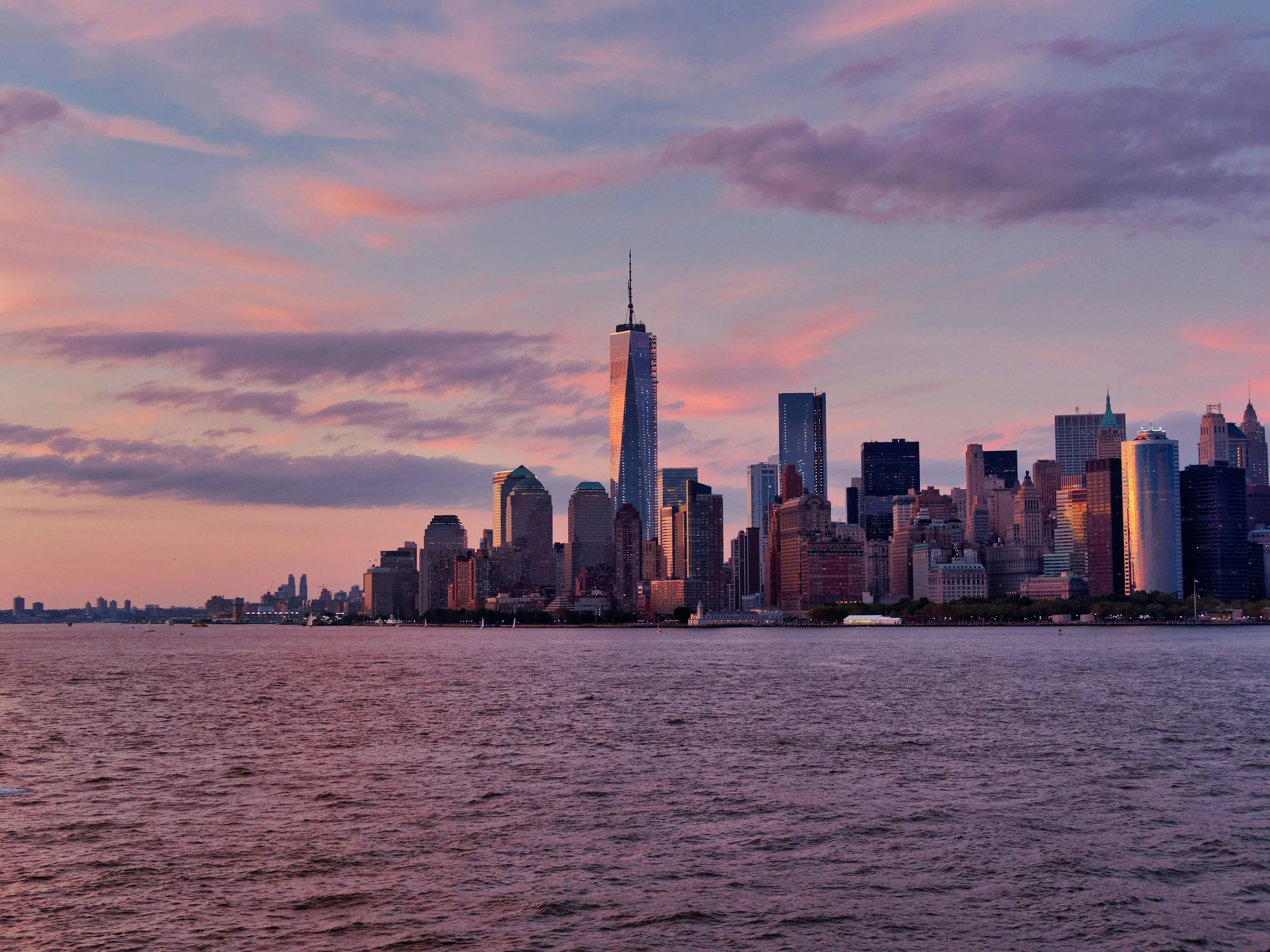 New York City skyline at sunset, with tall skyscrapers including One World Trade Center, viewed across the water.