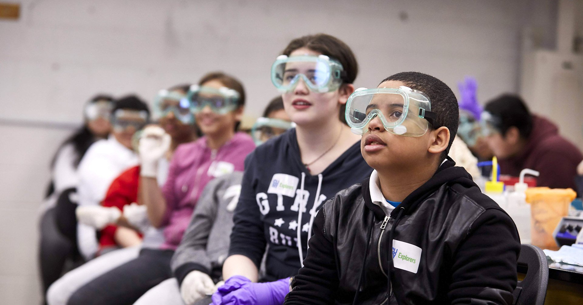 Children sitting in a classroom wearing safety goggles and gloves, participating in a science activity.