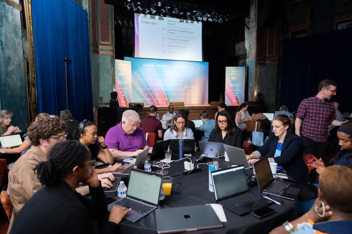 People working on laptops at a round table during a conference or workshop in an ornate room with blue curtains, with a stage and large screen in the background.