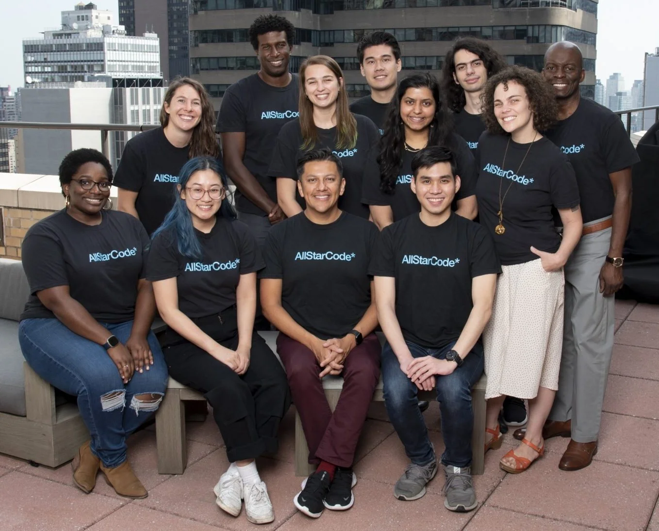 Group of diverse people in black T-shirts with 'AllStarCode' printed on them, smiling, on a rooftop with city buildings in the background.