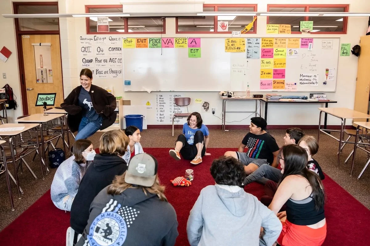 A group of children and two adults sitting in a circle on a red carpet in a classroom, with a whiteboard and classroom posters behind them. One child stands nearby, smiling.