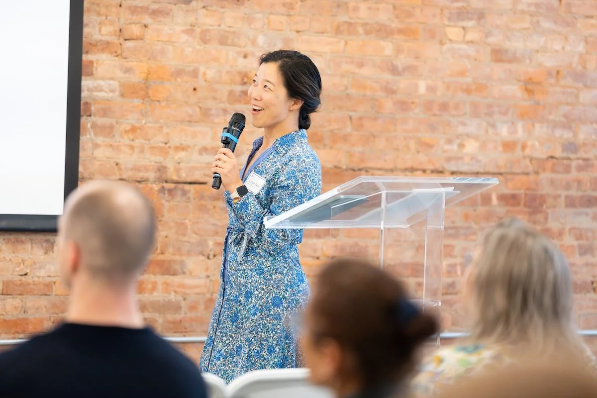 A woman in a blue floral dress giving a presentation with a microphone in front of a brick wall, audience members in the foreground.