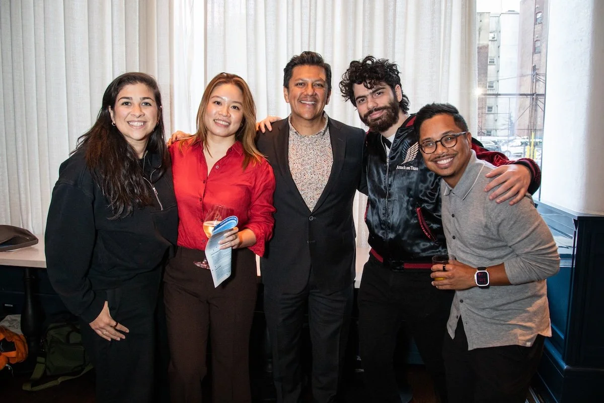 Six diverse people smiling and posing together indoors, with city buildings visible through a window in the background.