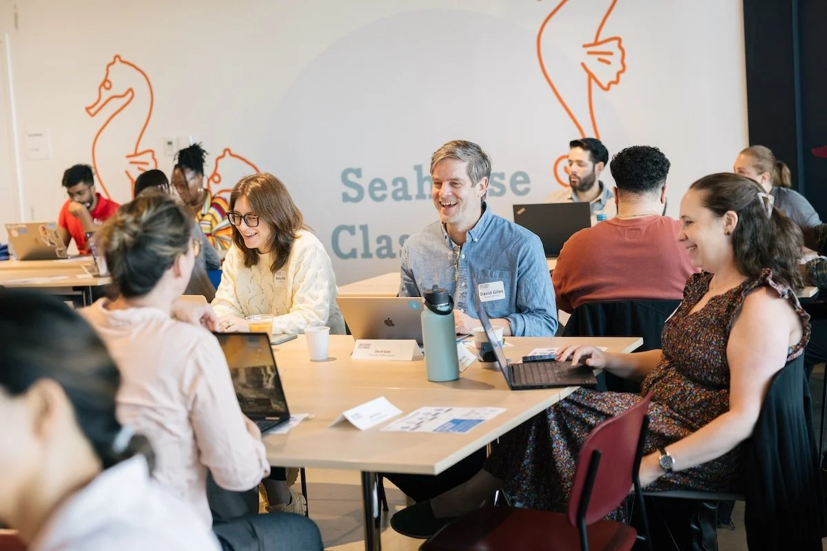 Group of diverse people attending a workshop or conference, sitting at tables with laptops and drinks, engaging in conversation and smiling, with a poster in the background that says 'Seahorse Classroom' and features seahorse illustrations.