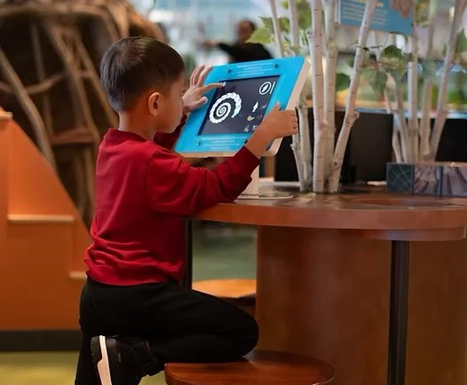 A young boy in a red shirt and black pants kneeling on the floor, using a tablet device at a wooden table, with tall plants in the background.