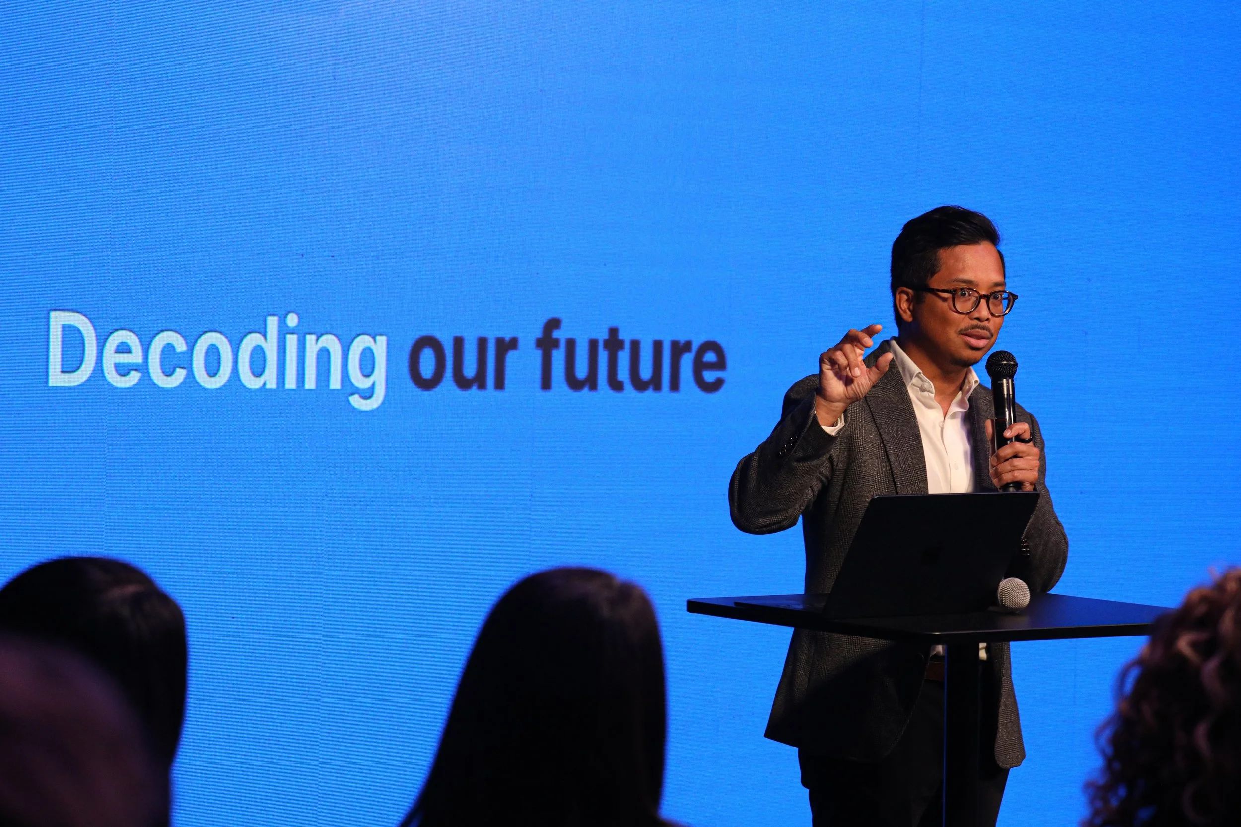 A man in glasses and a gray blazer giving a presentation on stage with a blue background. The slide behind him reads 'Decoding our future.' He is holding a microphone and standing near a laptop on a small table, with audience members visible in the foreground.