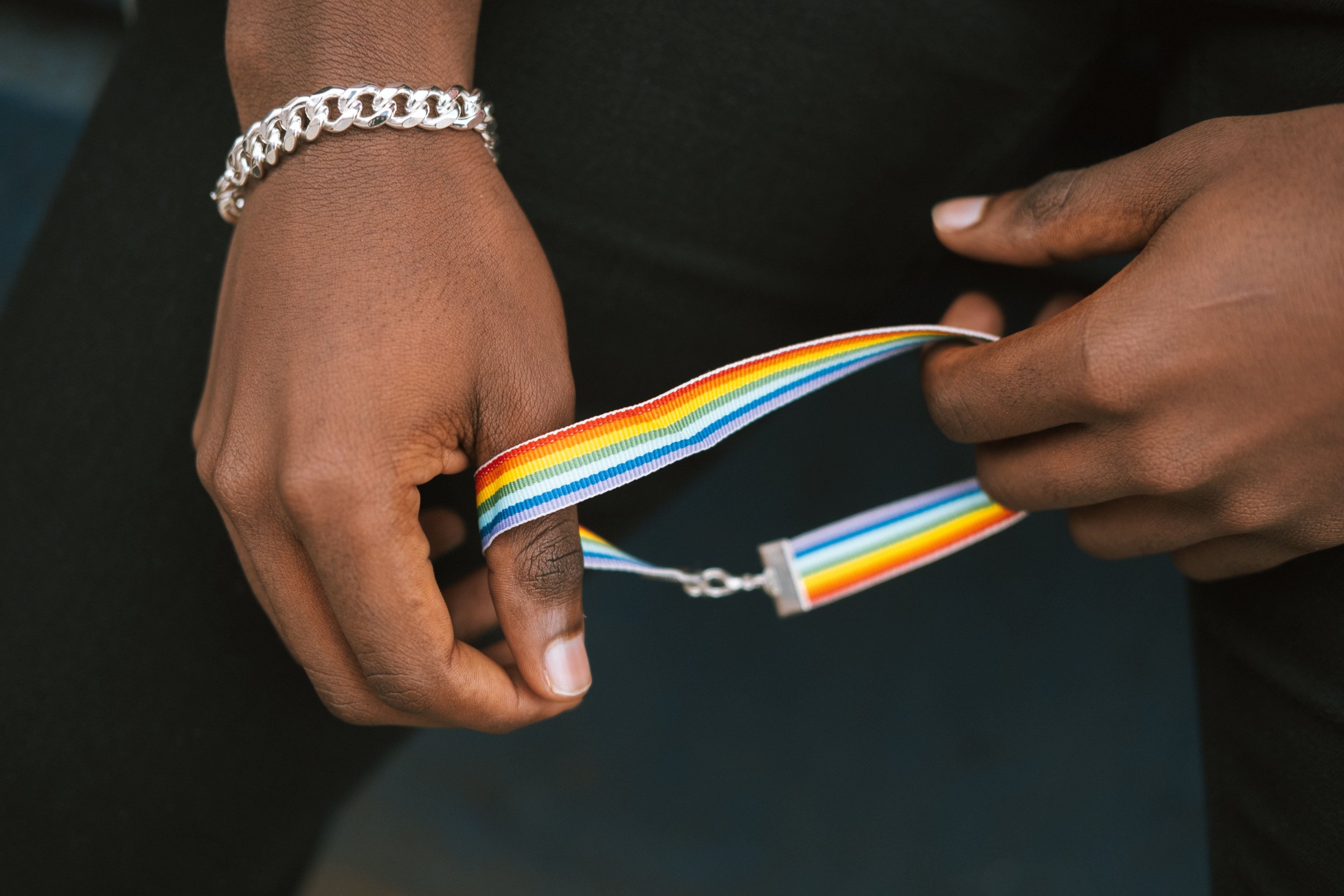 A black person's hands,  wearing a watch and bracelet holds a rainbow flag strap, symbolizing LGBTQ+ pride. Credit: Anete Lusina