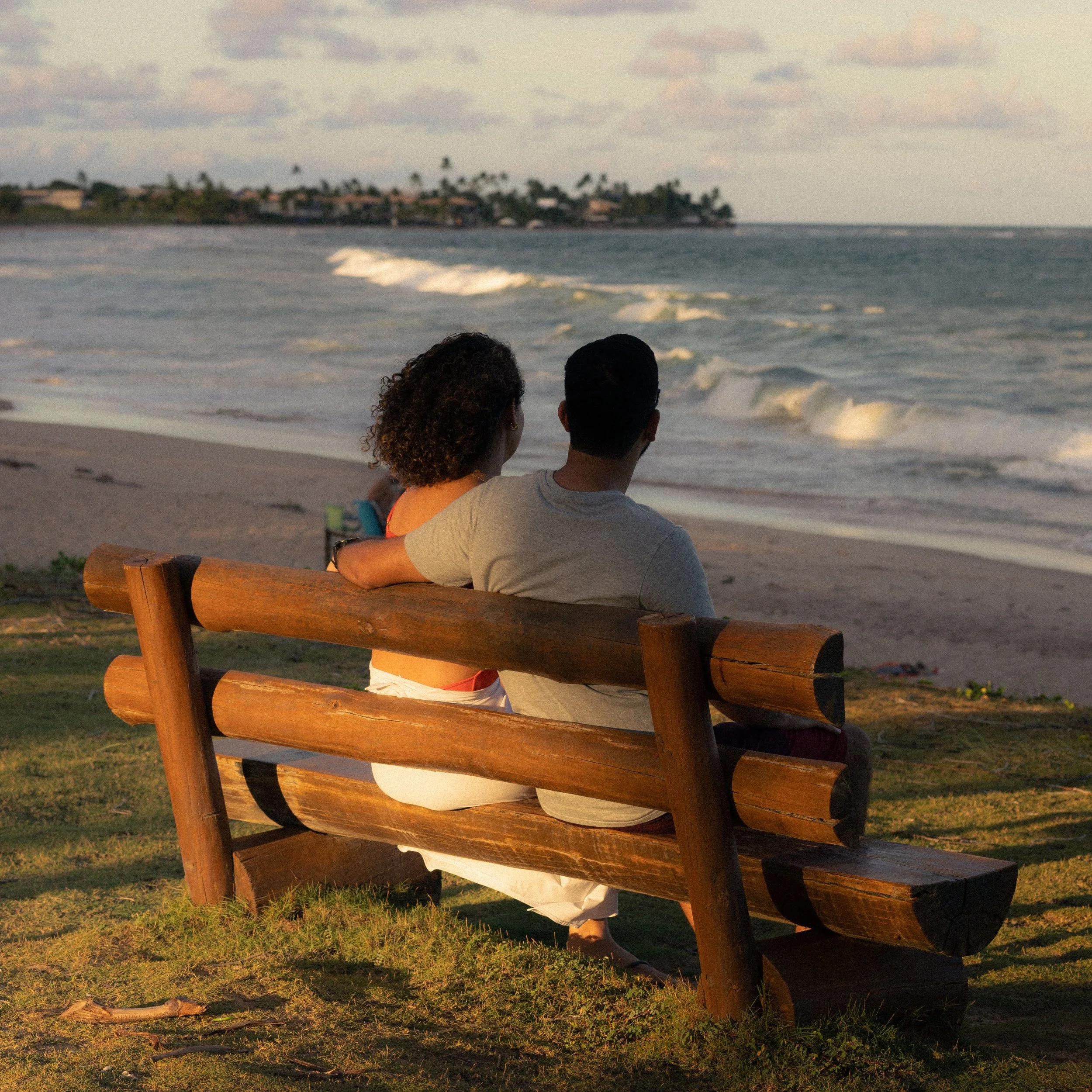 Couple facing away, embracing on a wooden bench overlooking the ocean at sunset. Credit: Alef Morais