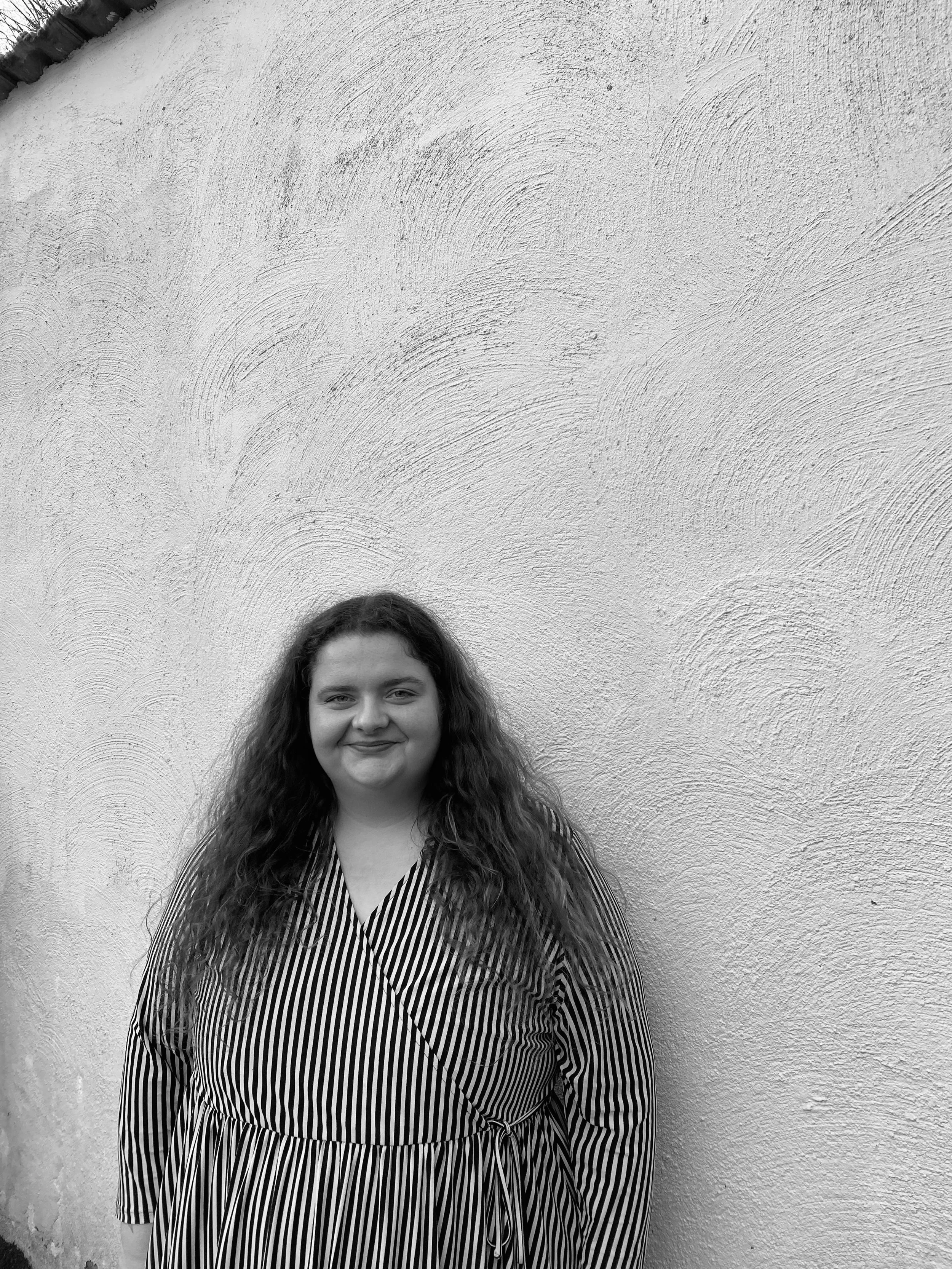 Junge Frau mit langen, gewellten Haaren, die vor einer weißen Wand mit struktureller Oberfläche steht, trägt ein gestreiftes Kleid.