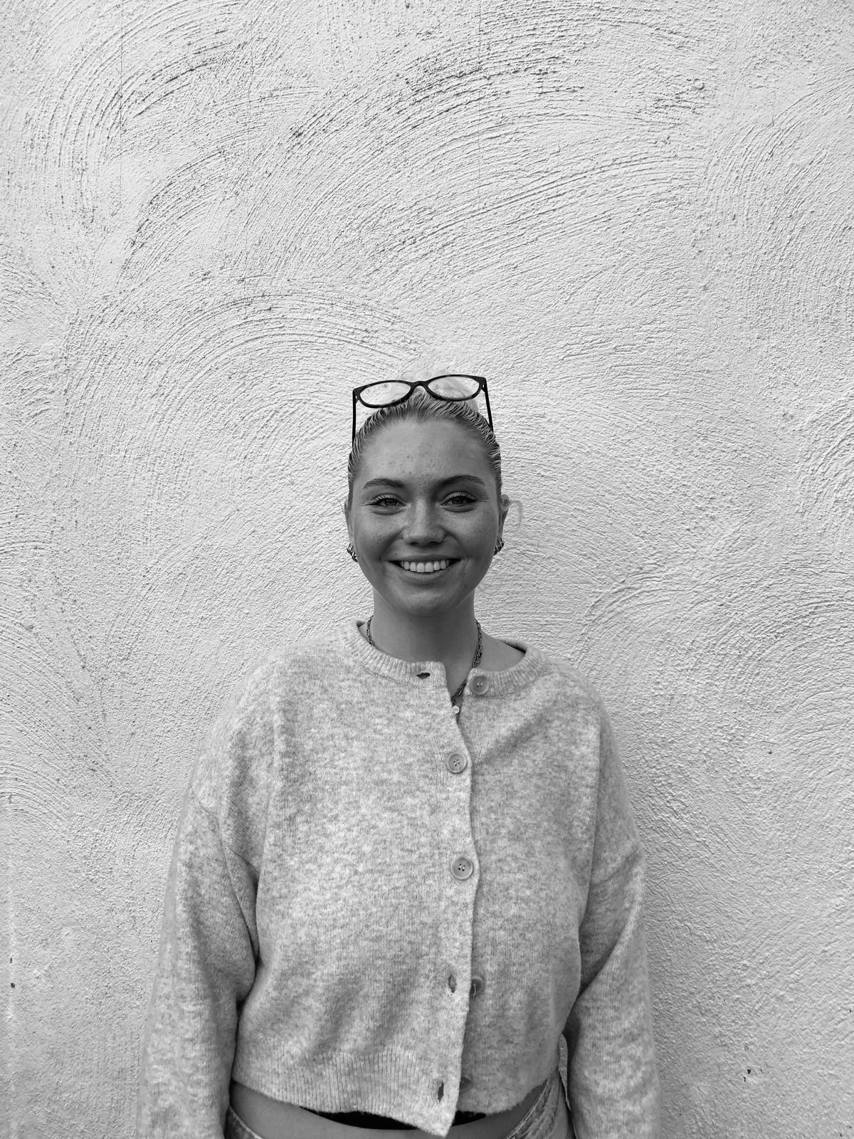 Junge Frau mit Brille auf dem Kopf, hat braunes Haar, trägt einen karierten Pullover und steht vor einer weißen Wand.