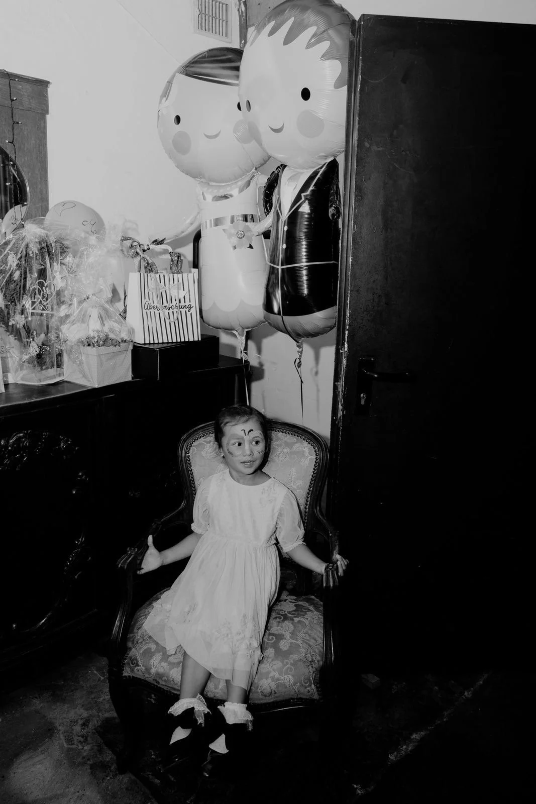Ein junges Mädchen im weißen Kleid sitzt in einem verzierten Sessel, trägt Clown-Schminke und hat eine eyebrows und Nasen-Make-up. Im Hintergrund sind heliumgefüllte Ballons, darunter Ballons, die wie eine Person in einem Anzug aussehen, und Geschenkartikel auf einem Tisch.
