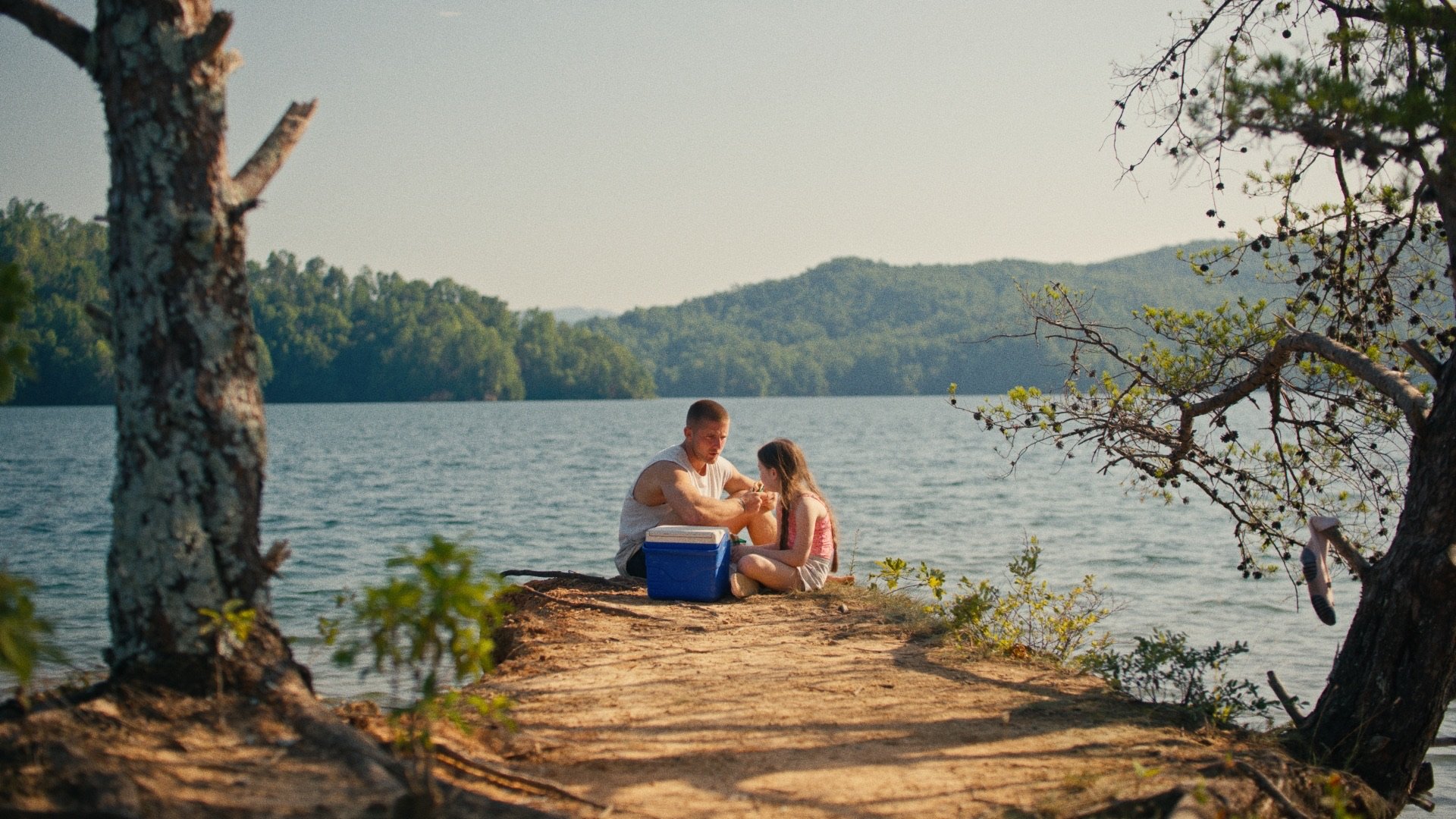 A man and a young girl sitting on a dirt path by a lake, surrounded by trees, with mountains in the background, enjoying a moment together.
