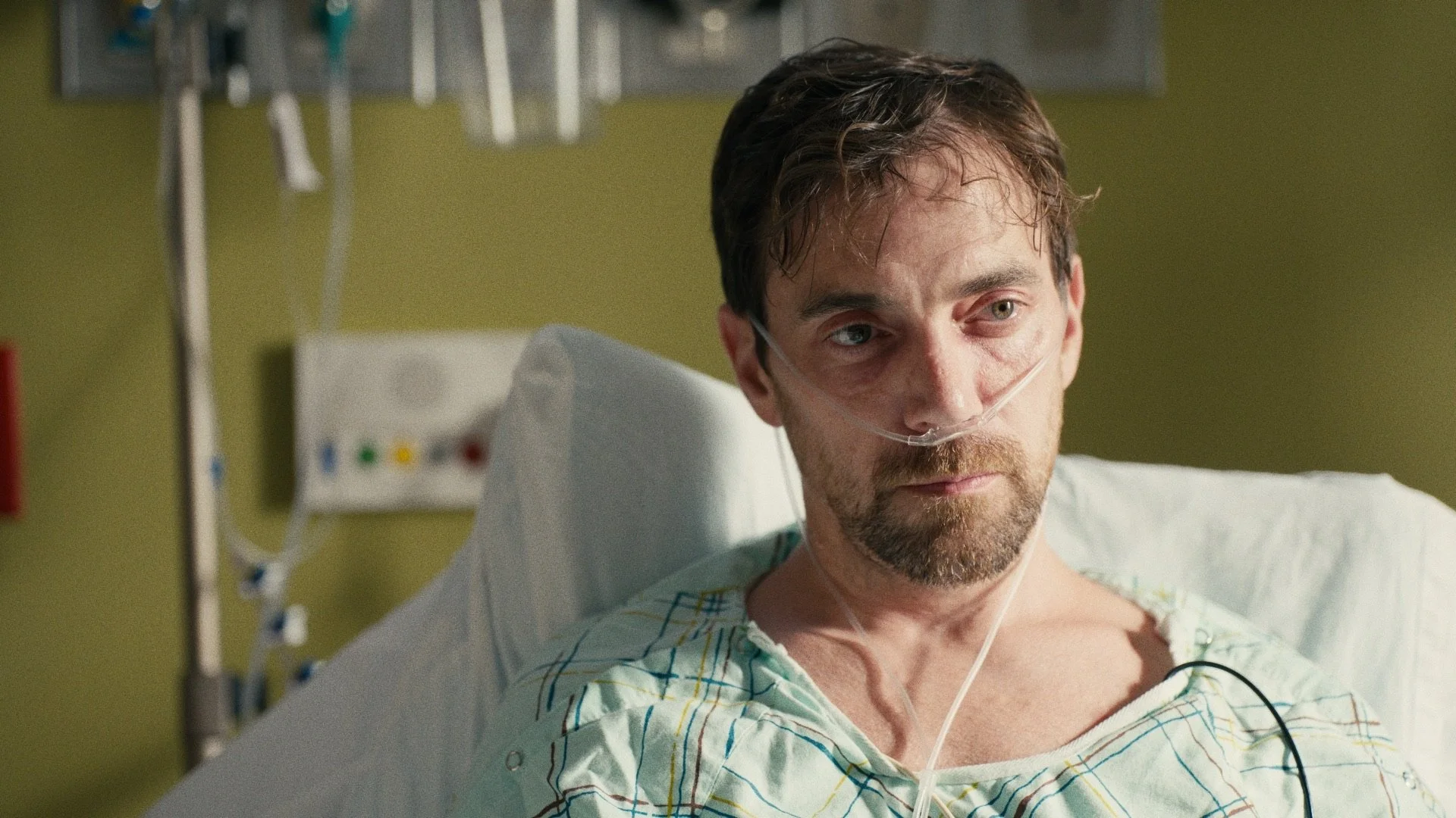 A man in a hospital bed wearing a hospital gown with a nasal cannula delivering oxygen, looking concerned and gazing into the distance.