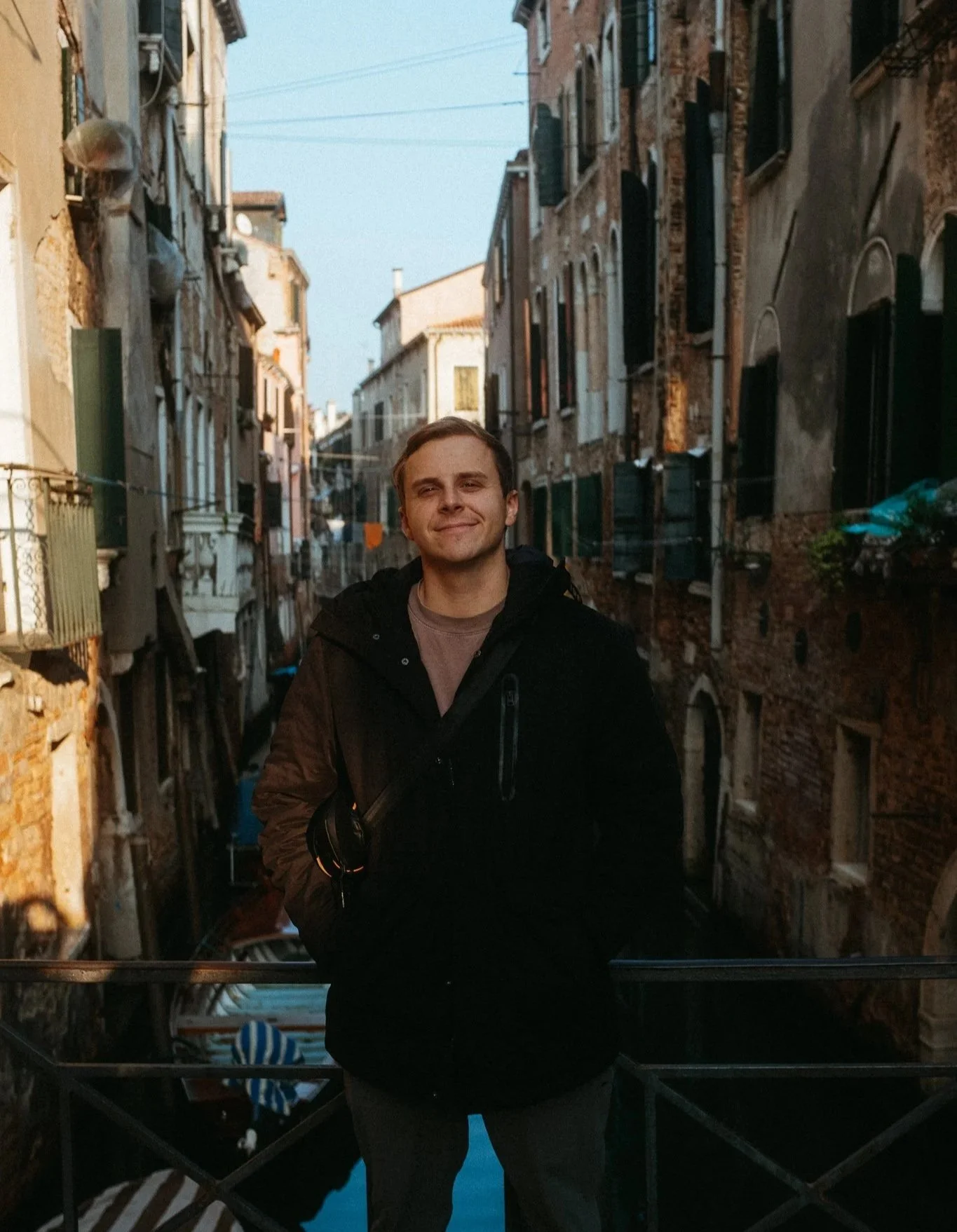 A young man smiling and standing on a bridge over a canal in Venice, Italy, during daylight.