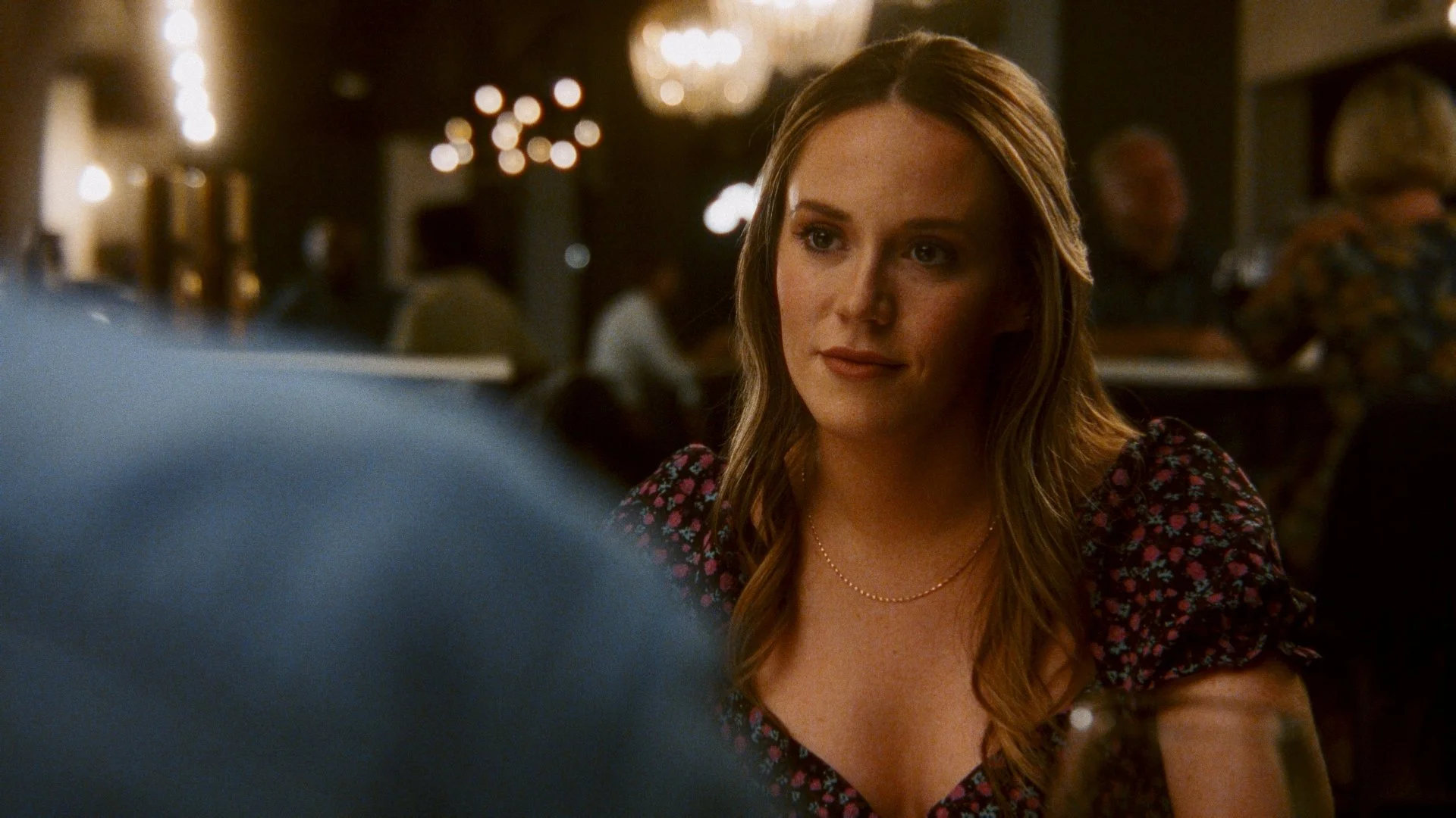 A woman with long, wavy red hair sitting at a table in a dimly lit restaurant, wearing a floral dress and a pearl necklace, engaged in conversation.