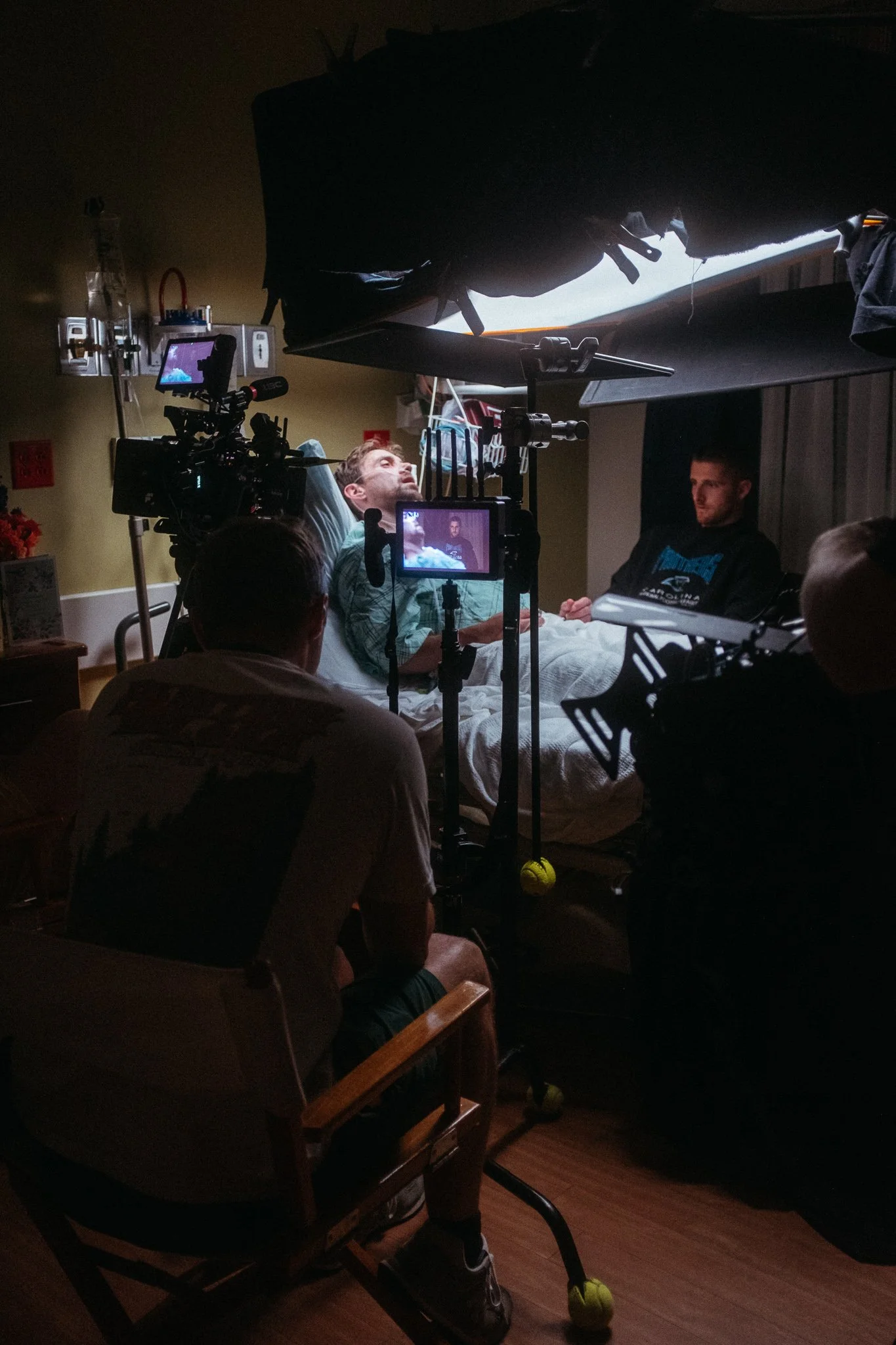 Behind the scenes of a medical procedure filming, with a man in a hospital bed, surrounded by film crew and equipment.