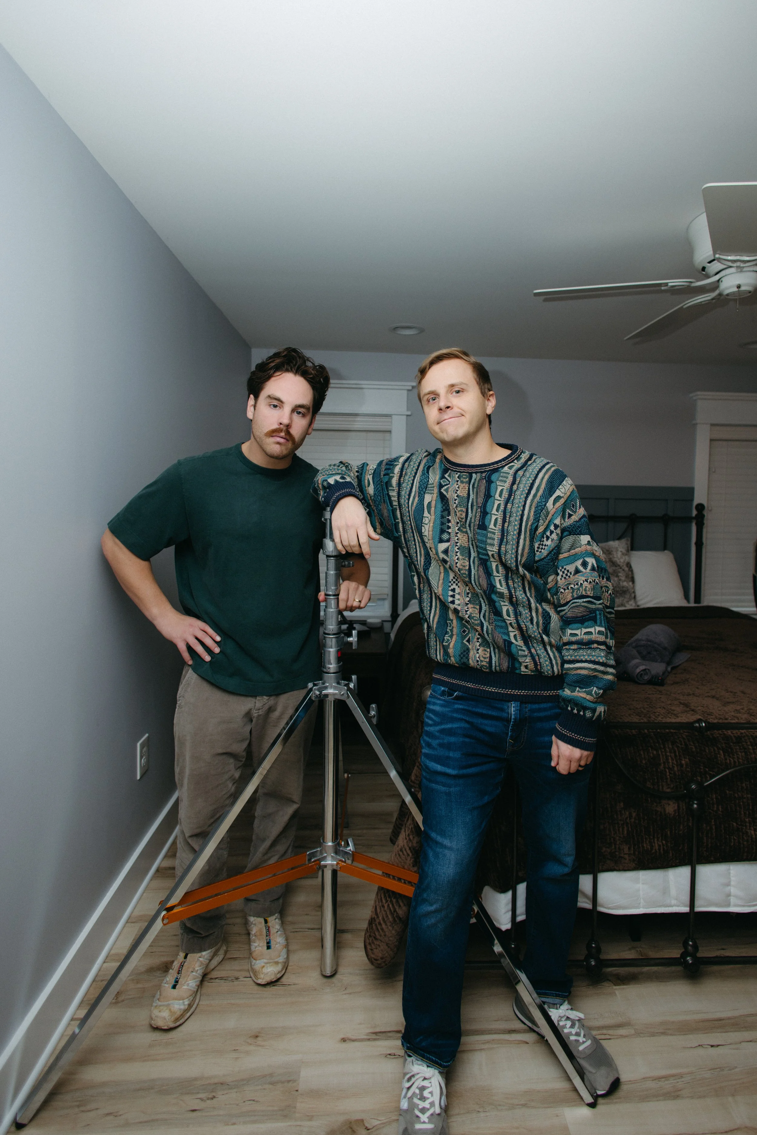 Two men standing in a bedroom, one leaning on a tripod with a camera or equipment, both looking at the camera, in a cozy bedroom with beds and rolled towels in the background.