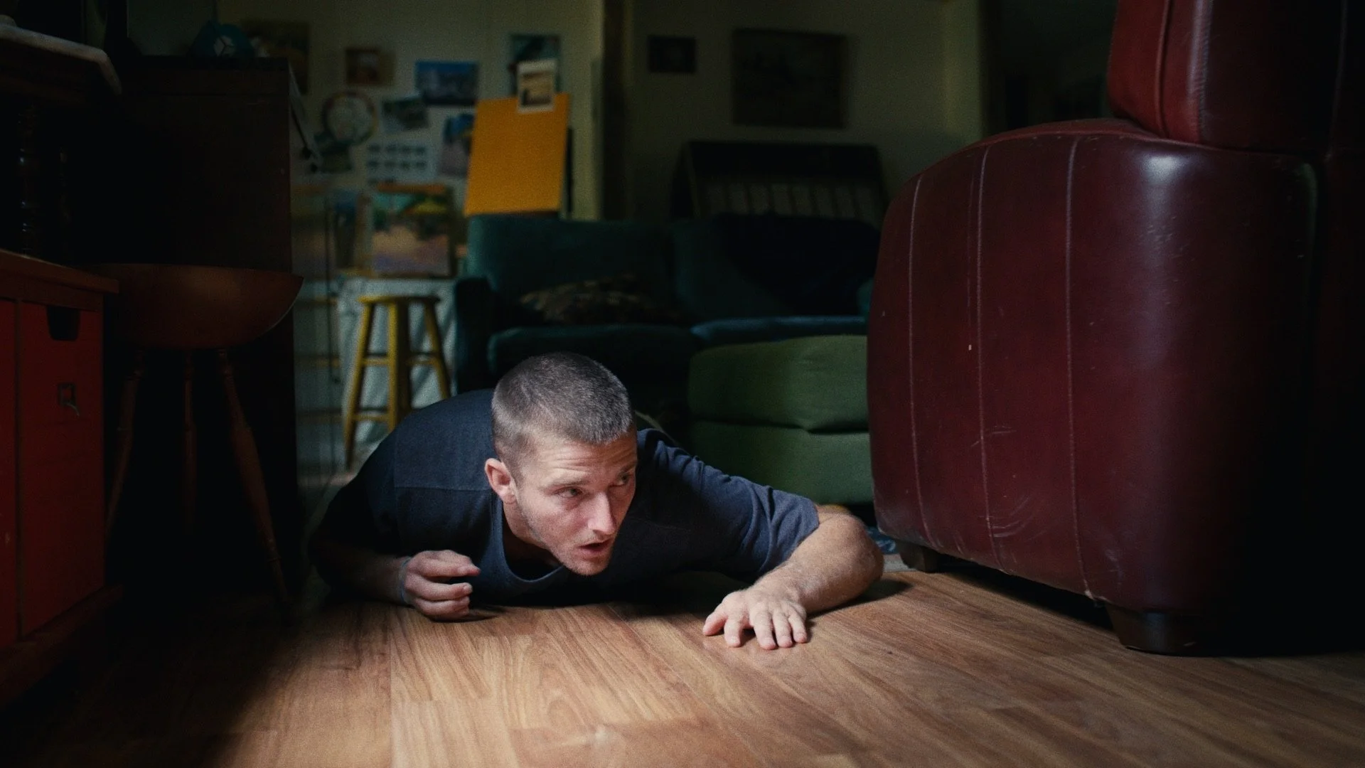 A man crawling on the floor in a dimly lit room, looking underneath a red leather sofa.