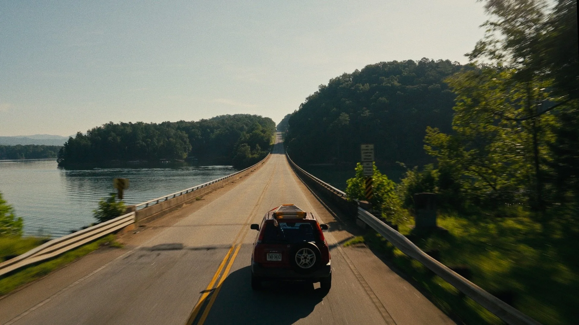 A red SUV driving on a narrow bridge over a body of water with green trees on both sides and mountains in the background, during daytime.
