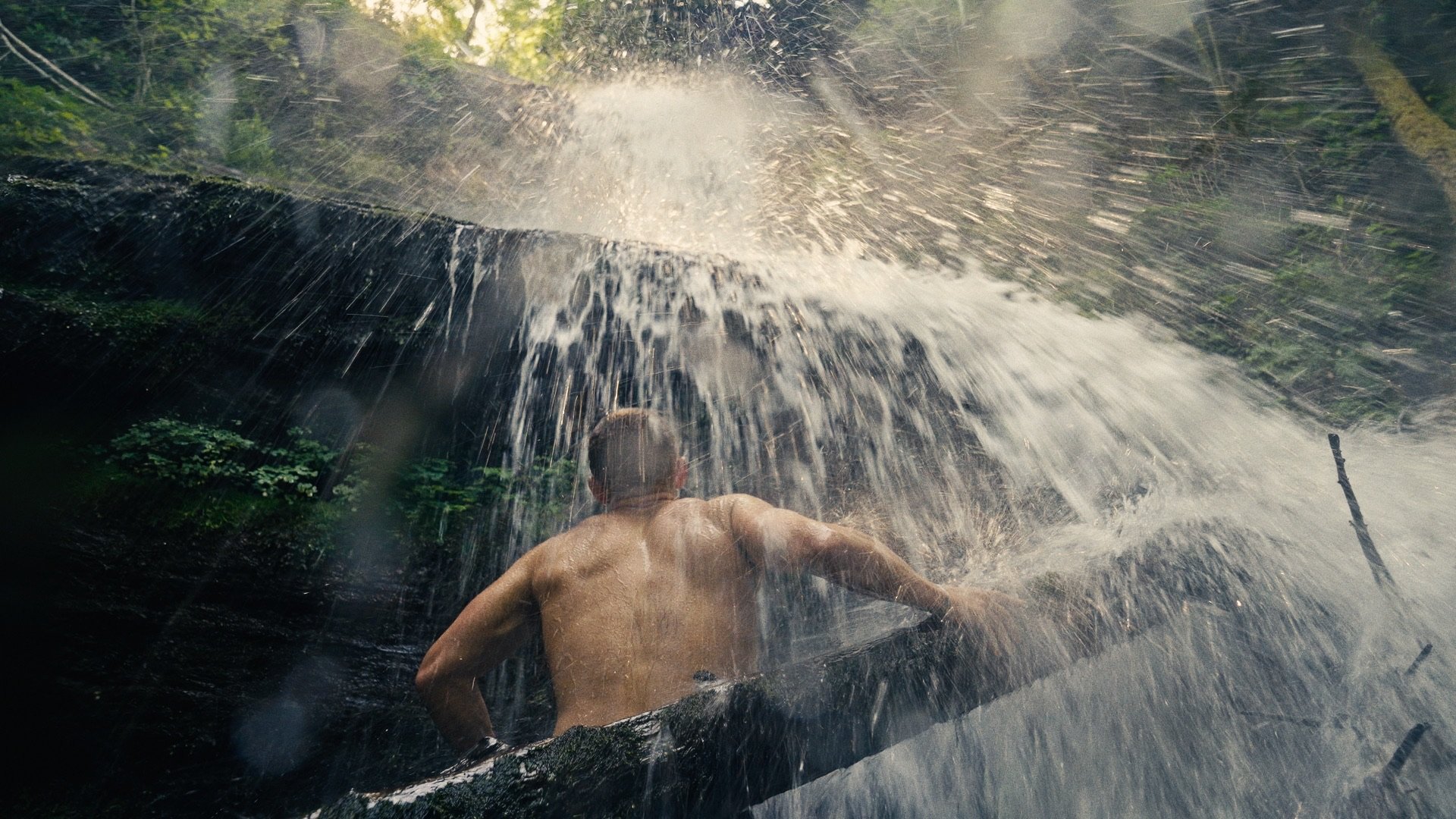 A person with a bare back climbing up a wet, mossy rock face under a waterfall in a forest.