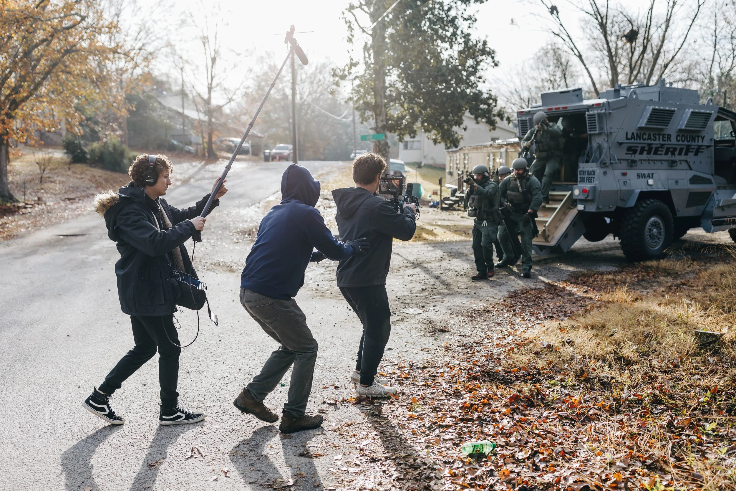 Film crew filming a police SWAT team entering a house with a SWAT vehicle parked outside in a suburban neighborhood.