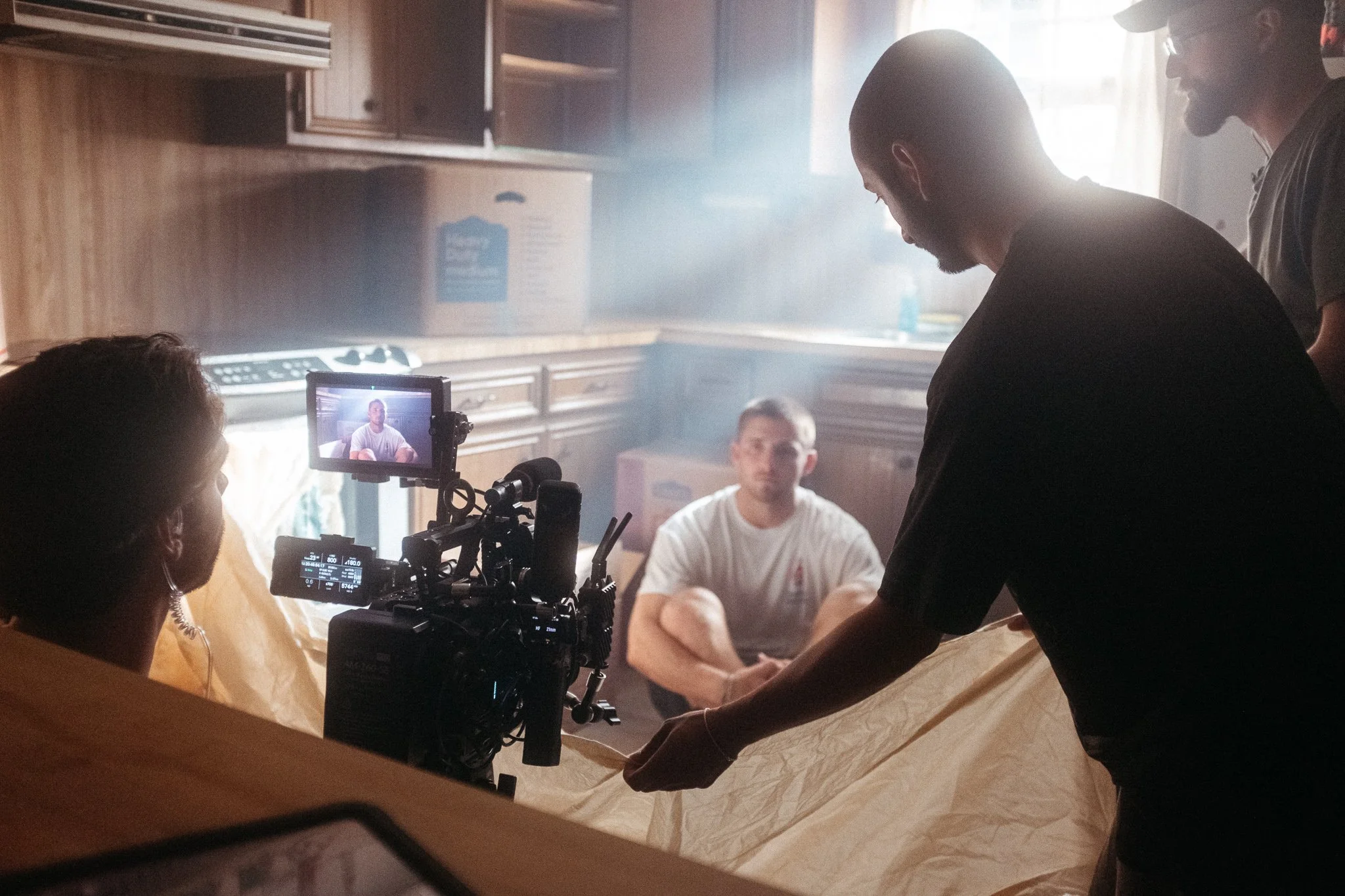 Film crew members filming a man sitting on the floor in a kitchen with wooden cabinets, using professional camera equipment and a backdrop of natural light.