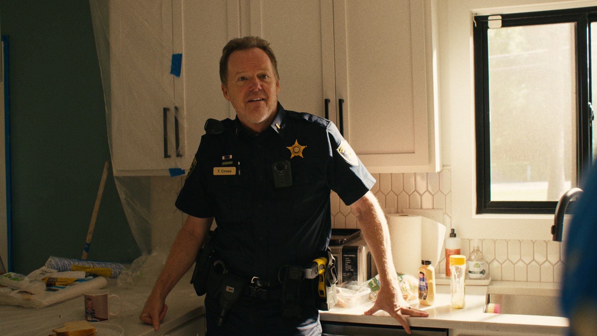 A police officer in uniform standing in a kitchen with various items on the counter, and a window in the background.