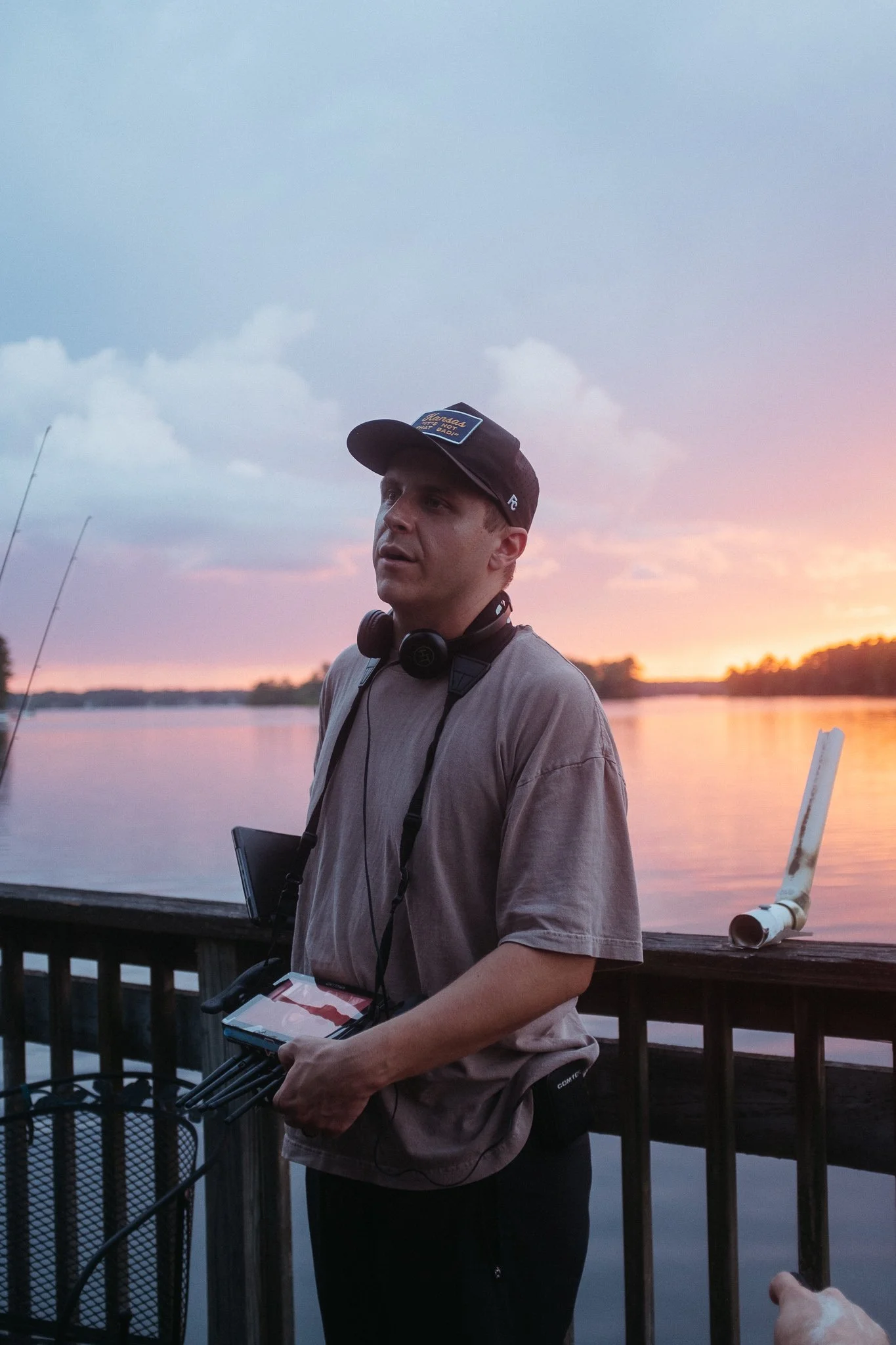 Man with headphones holding fishing equipment on a dock during sunset by a lake.
