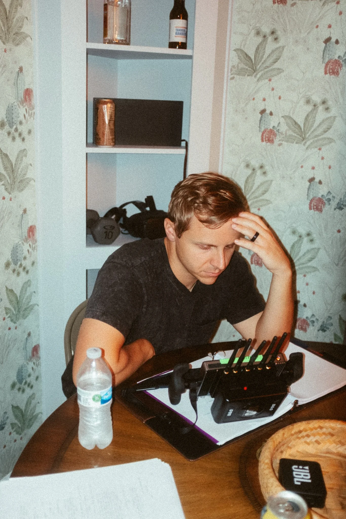 A man sitting at a dining table, looking down with a concerned or focused expression, with a water bottle, some papers, and a Wi-Fi router in front of him. Background includes a built-in shelf with some items, a floral-patterned wall, and a backpack.