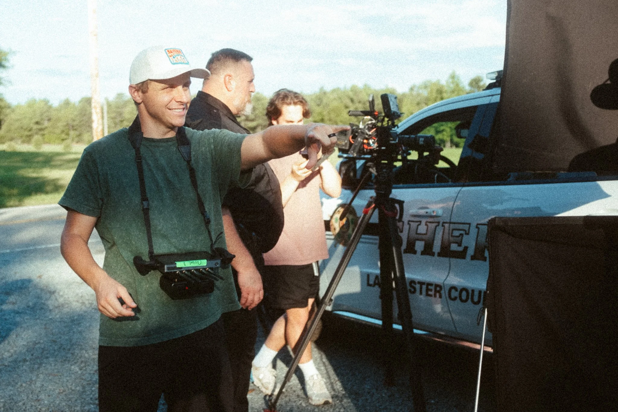 A person with a camera crew filming a police car on the side of the road during daytime.