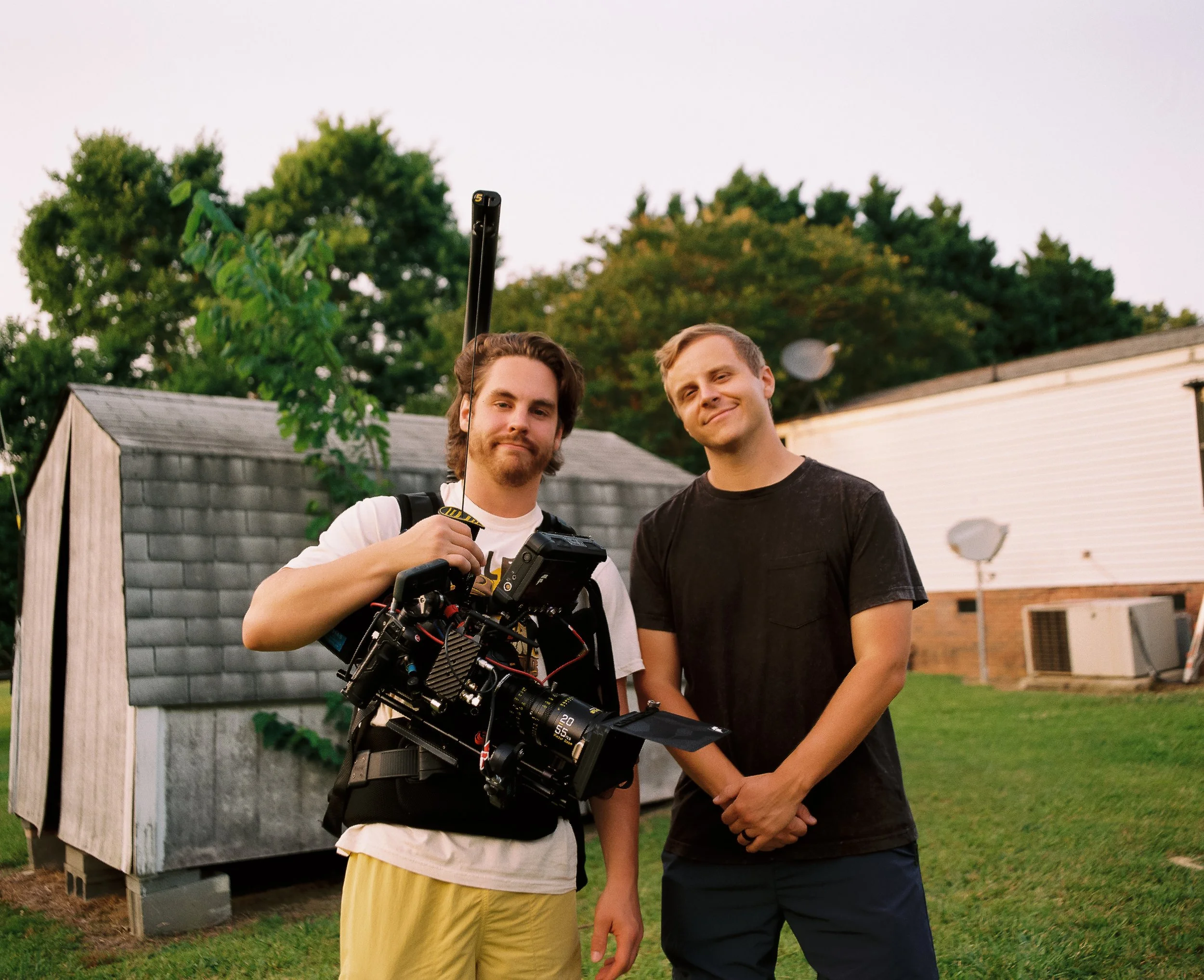 Two young men standing outside on grass, one holding a camera and equipment, the other smiling, with trees and buildings in the background during dusk.