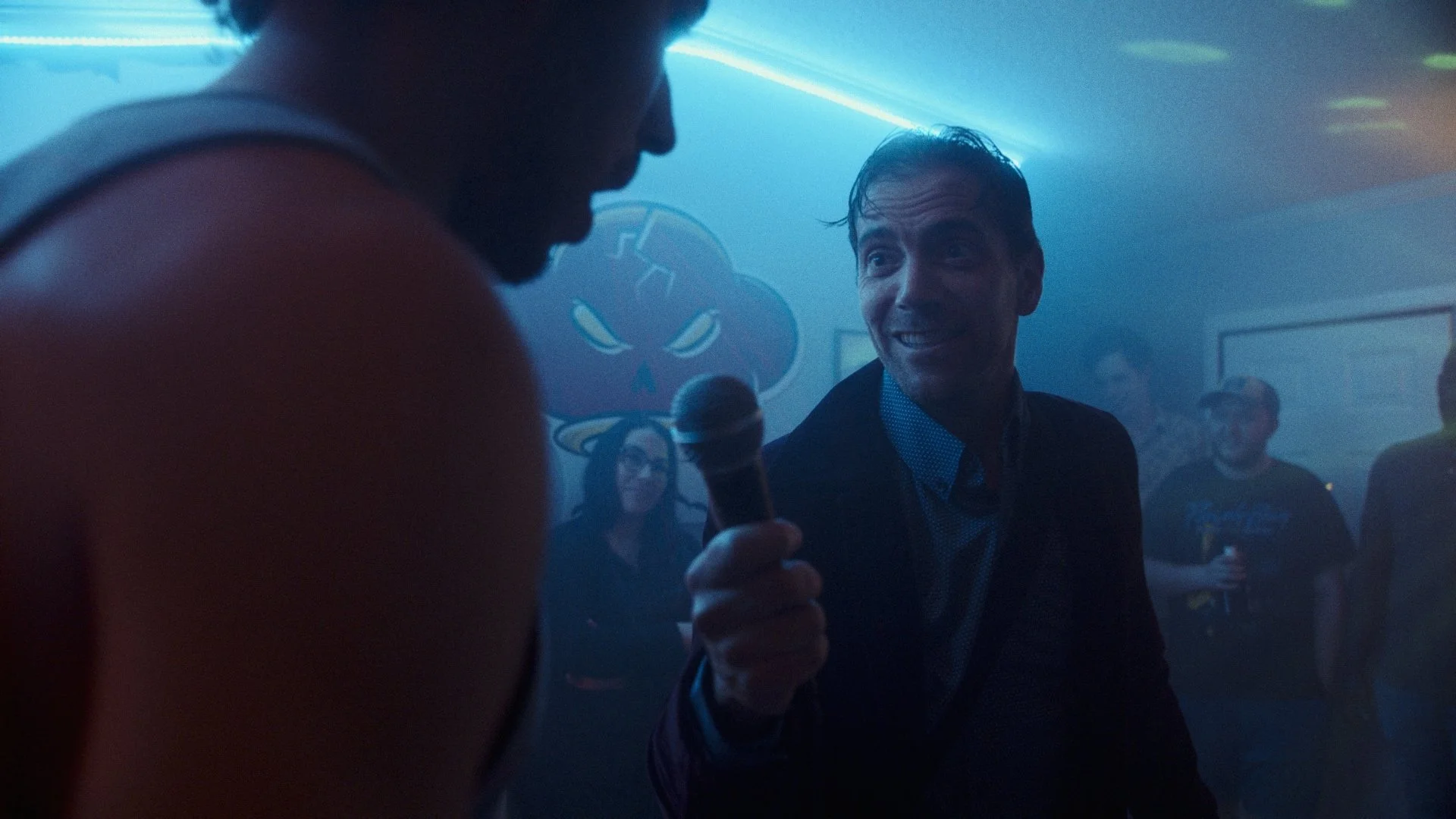 People in a dimly lit bar or club, with a person holding a microphone, and others watching. Background features neon lighting and a logo with a skull and hat.