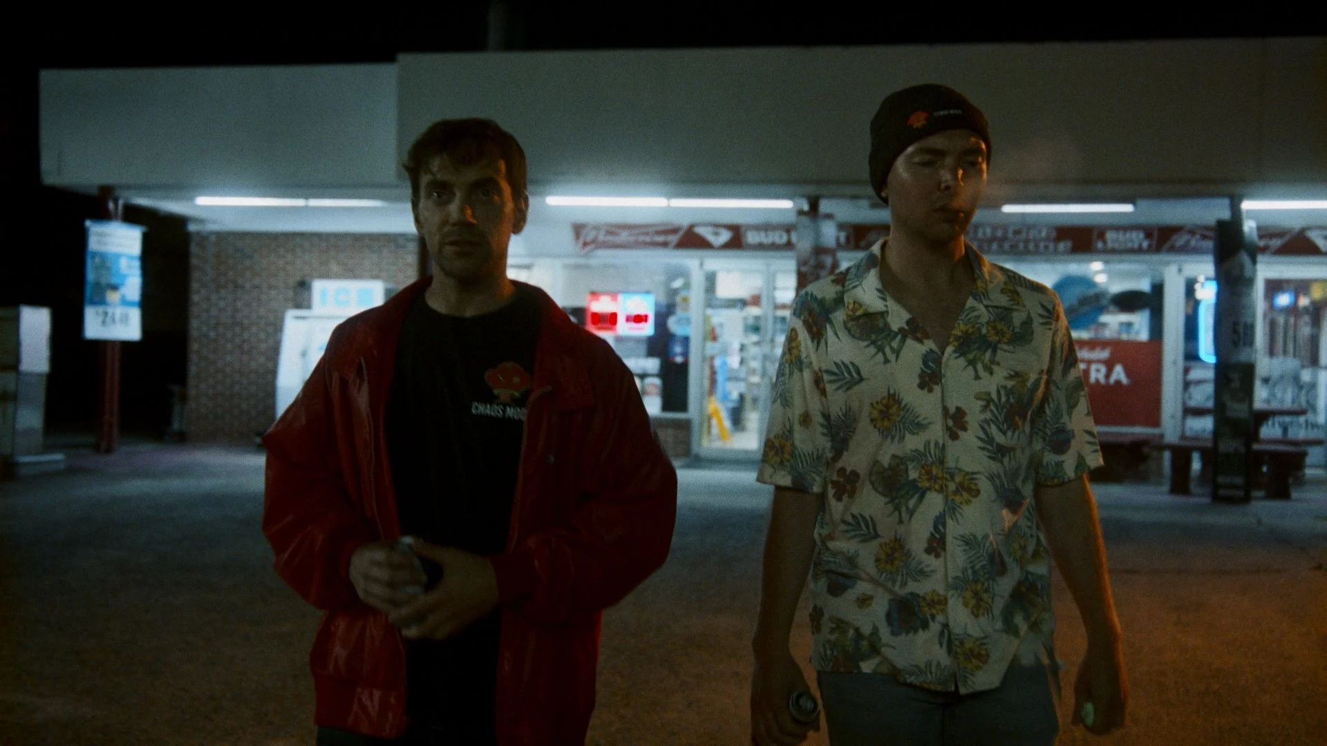 Two men walking outside a convenience store at night, one wearing a red jacket and the other in a floral shirt and black beanie.