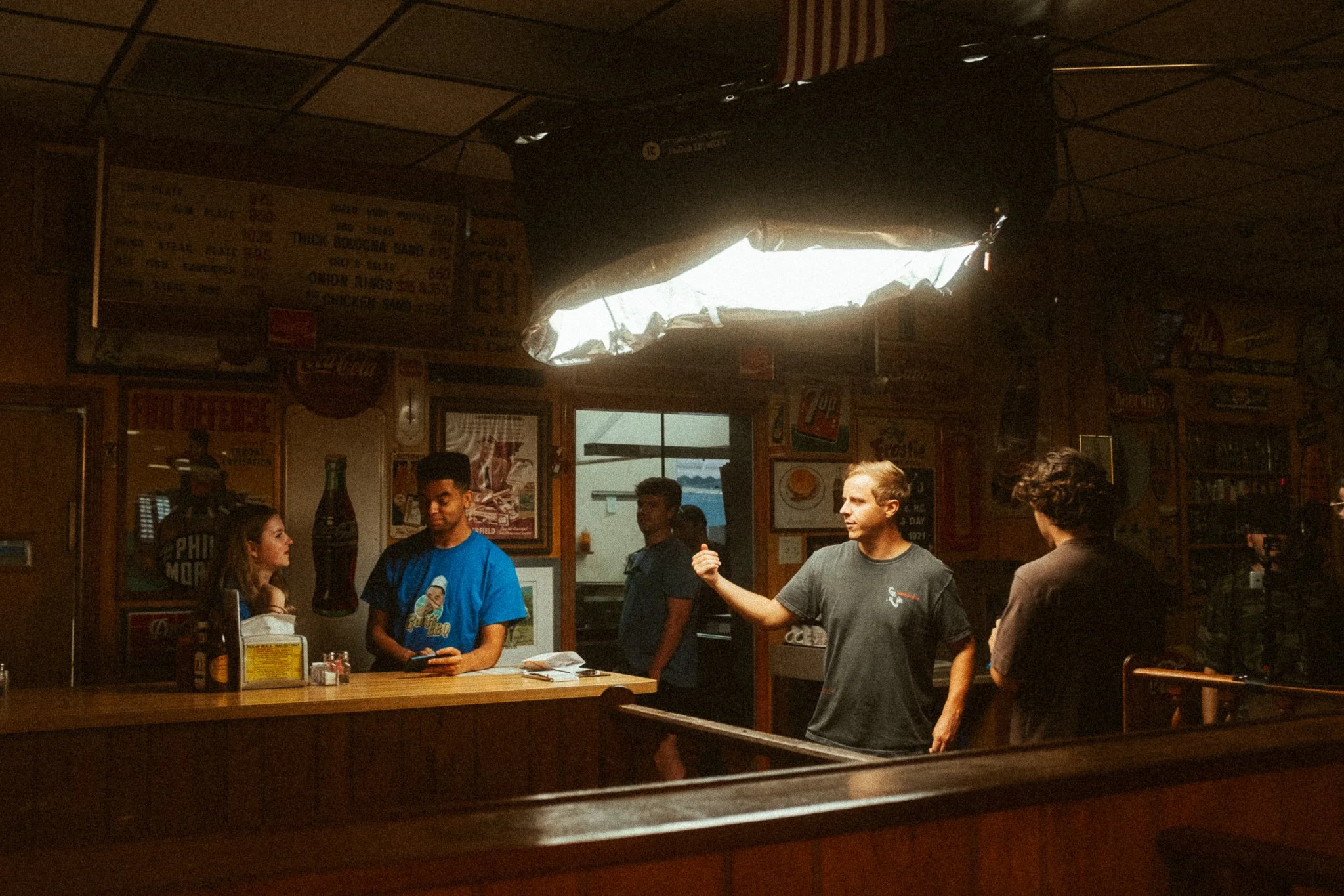 Inside a restaurant with four people, including a woman at the counter and three men, one gesturing with his hand. The dimly lit space has wood-paneled decor and nostalgic advertisements on the walls.