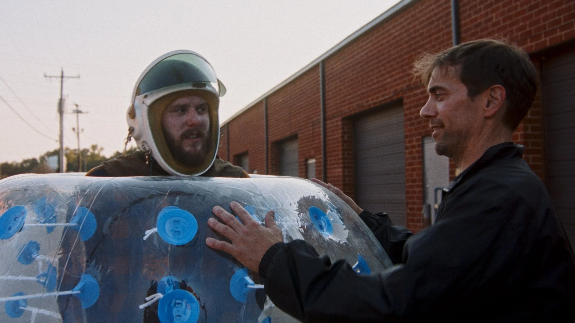Two men, one in a helmet and the other in a black jacket, interact near a transparent vehicle covered with blue suction cup-like devices, outside a brick building.