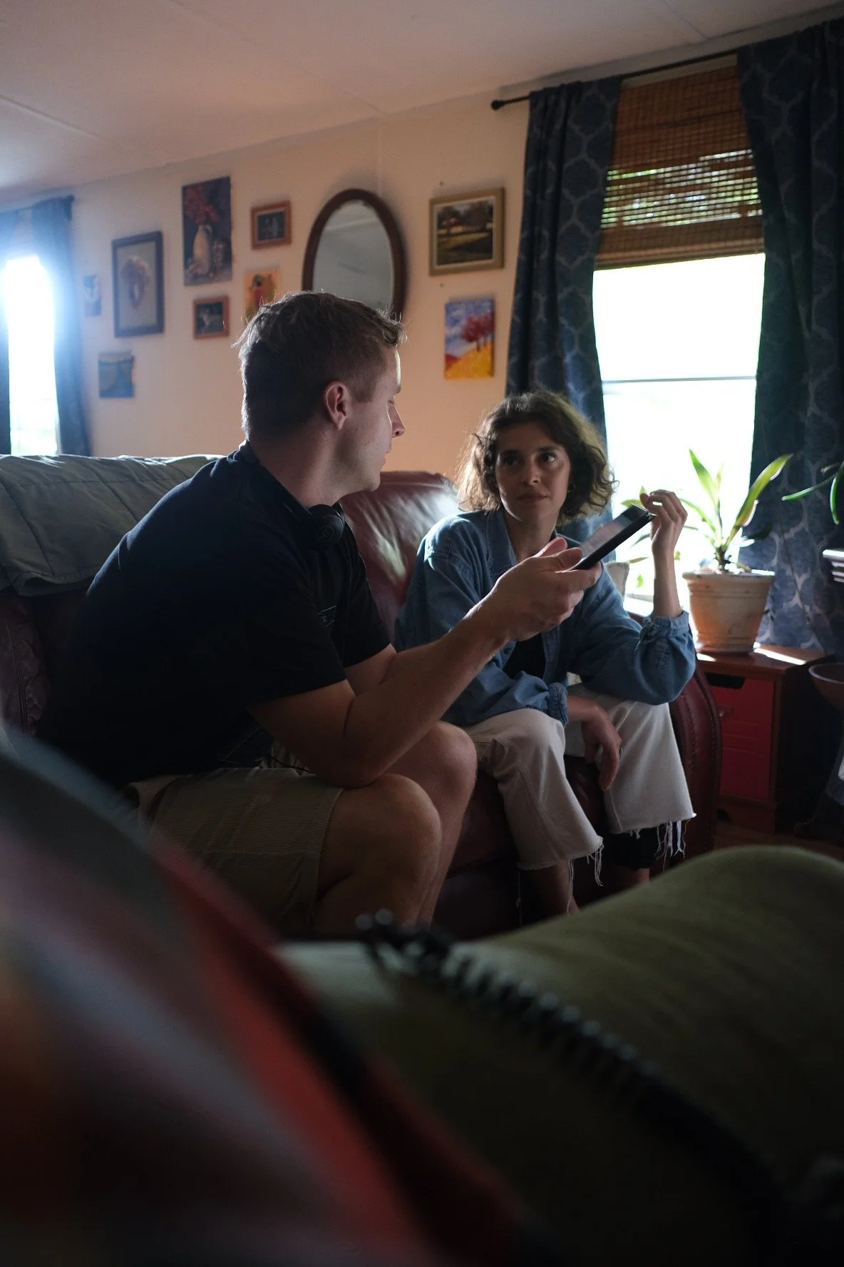 Two people sitting on a brown leather couch in a living room having a conversation, with a man holding a smartphone and a woman listening attentively.