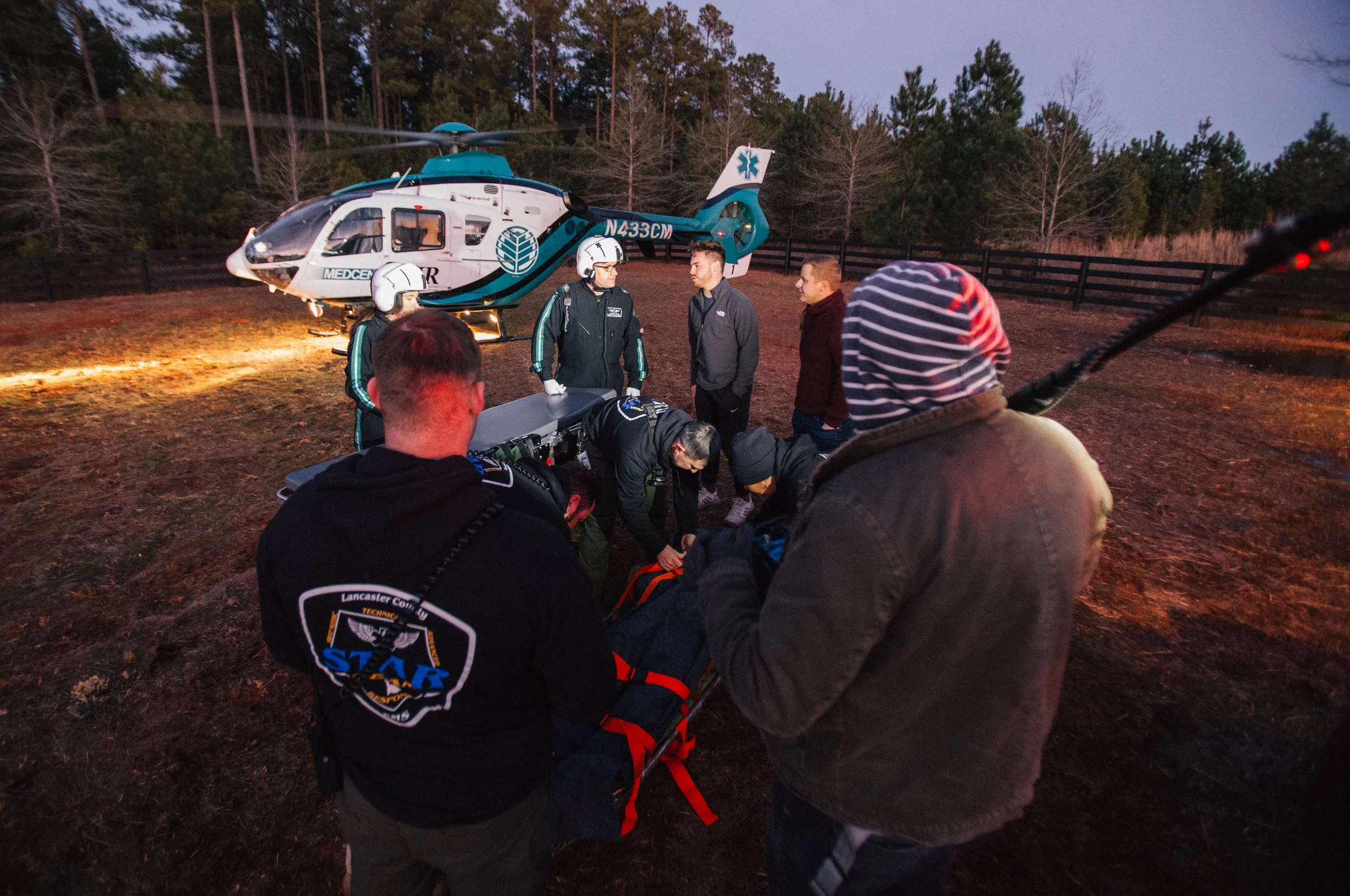 Paramedics and emergency responders attending to a person on a stretcher outdoors at dusk, with a helicopter in the background.