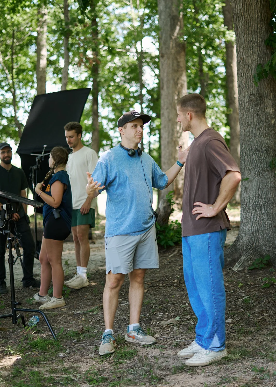 Two men are having a conversation on a film set in a wooded area, with film crew and equipment in the background.