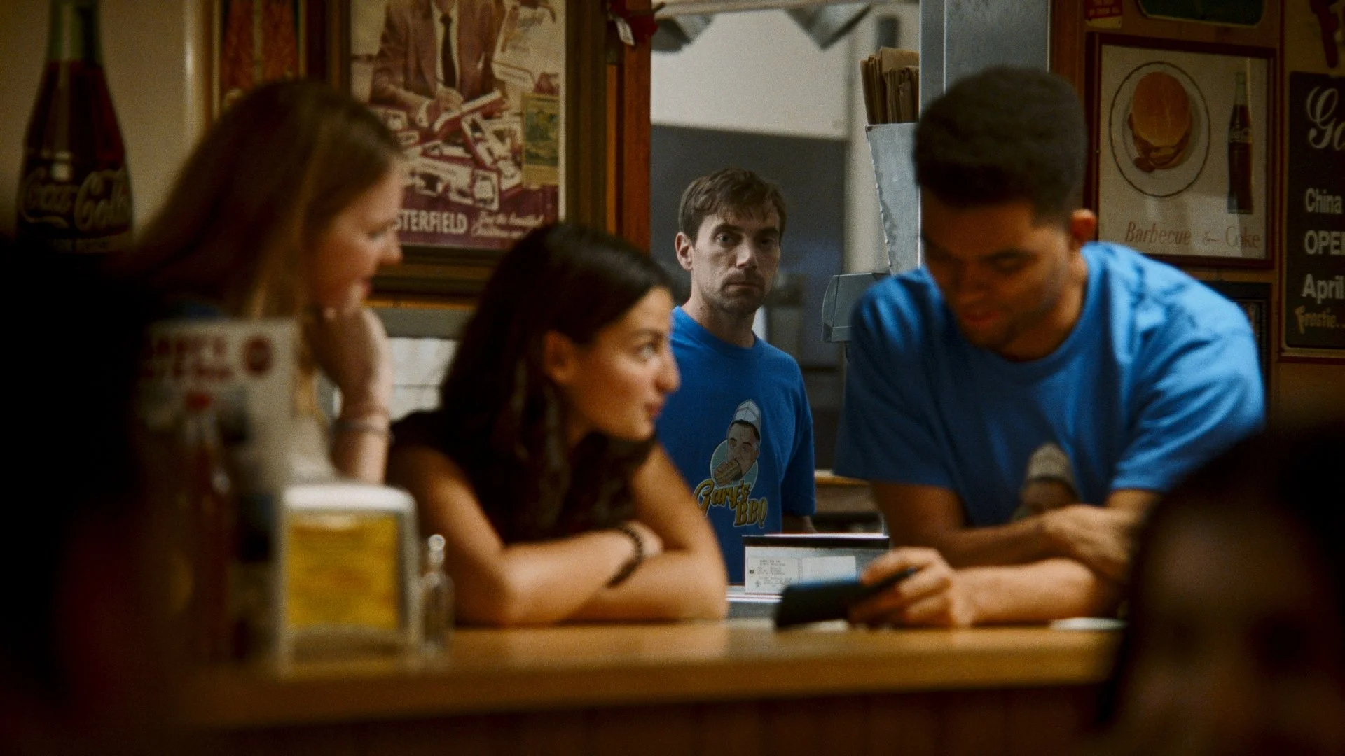 Four people sitting at a bar or restaurant, with one man in a blue T-shirt looking directly at the camera, while the others are engaged with their phones or in conversation.
