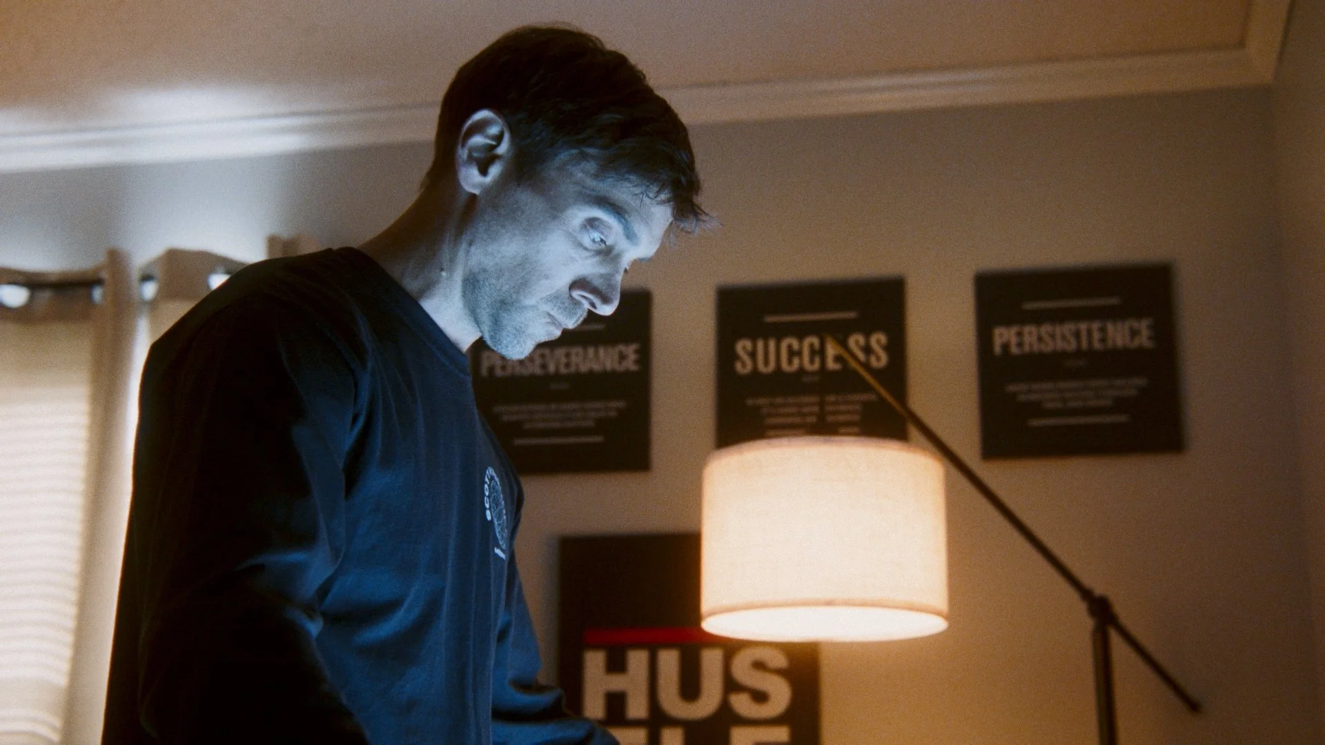 A man with short dark hair and a beard looking down, illuminated by a lamp, with motivational posters on the wall behind him reading 'PERSEVERANCE,' 'SUCCESS,' and 'PERSISTENCE'.