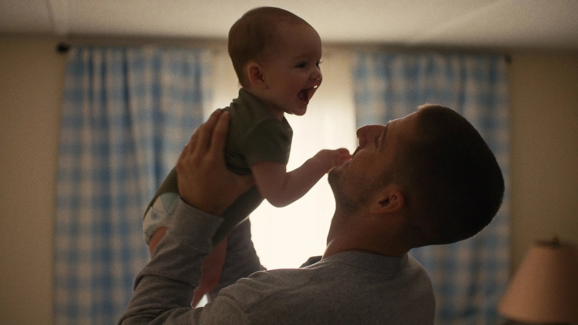 A father lifting his young child, both smiling and happy inside a warmly lit room.