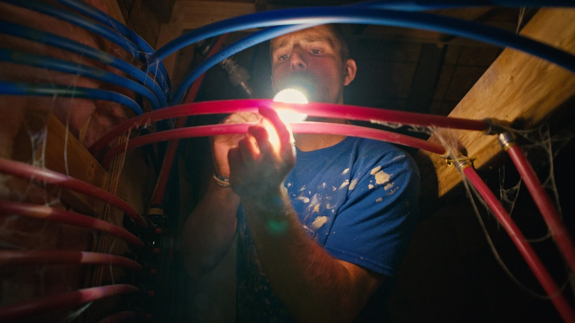 A man working on electrical wiring, using a flashlight, surrounded by bundled red and blue cables in an attic or crawl space.